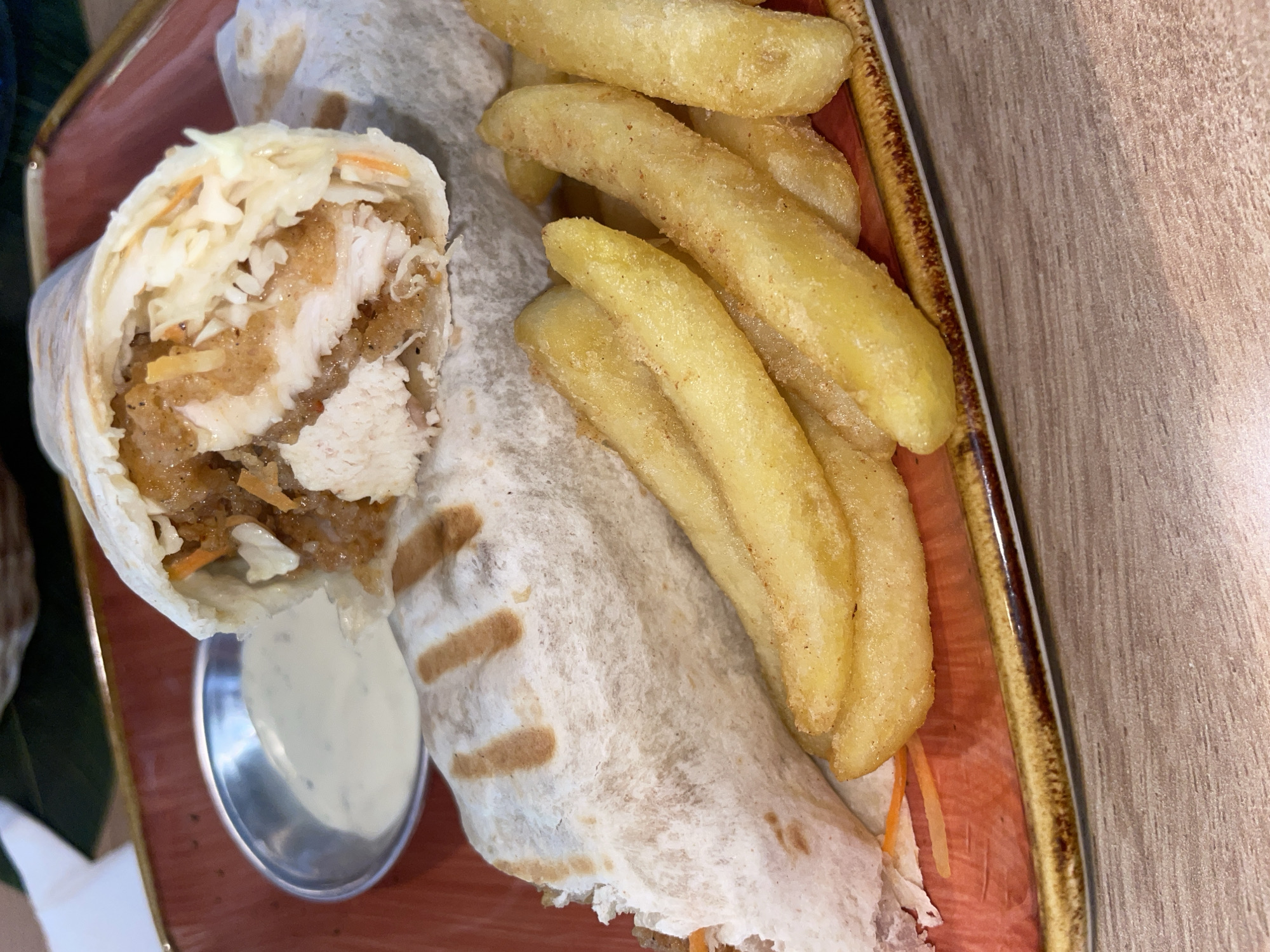 A close-up, top-down shot showcases a tightly wrapped chicken wrap and a side of french fries on a reddish-brown, textured ceramic plate. The wrap, partially opened, reveals a generous filling of shredded chicken, coleslaw, and what appears to be a sauce. The french fries are thick-cut and golden brown, seasoned with visible specks of seasoning. A small, silver dipping dish filled with a creamy white sauce is positioned to the left of the wrap. The plate rests on a light brown, wooden surface, suggesting a dining setting. The image is professionally lit, highlighting the textures and colors of the food. The scene does not provide any information about the city of Podgorica, Montenegro.