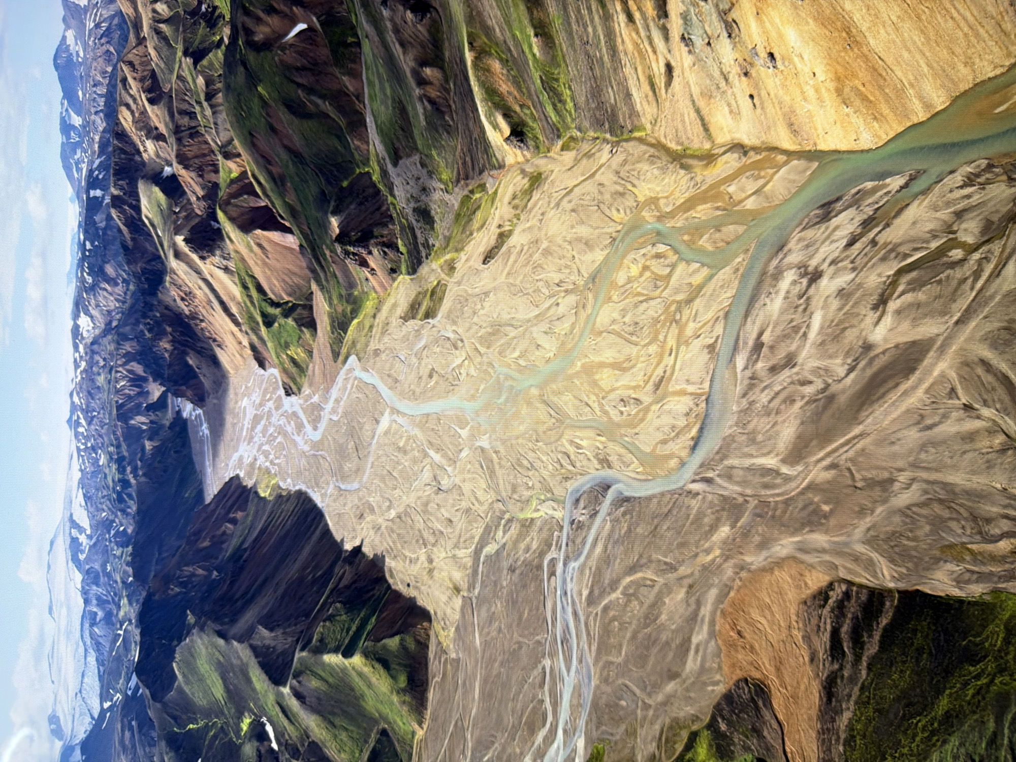 The image depicts an aerial view of a vast, braided river system flowing through a rugged, mountainous landscape. The river's channels are a pale, sandy color, creating intricate patterns across the valley floor. The surrounding mountains display a mix of earthy tones, ranging from tan and brown to green, with some patches of snow visible on the higher peaks in the background. The sky is blue with scattered clouds. The scene is a natural landscape, with no indication of any urban environment, including the city of Kyiv, Ukraine. There are no people, objects, or actions visible in the image.