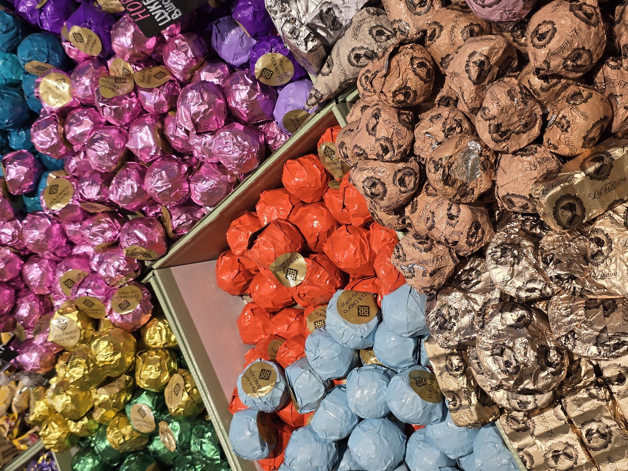This is a close-up, top-down shot of a colorful assortment of individually wrapped chocolates, likely displayed in a store or market. The chocolates are arranged in bins or sections, each section filled with candies wrapped in distinct foil colors. From left to right, there are sections of teal, pink, purple, yellow, green, orange, light blue, and gold/brown. Many of the wrappers have visible text. Some wrappers, particularly those in the pink, yellow, and teal sections, feature a gold circular sticker with the text "LUX CHOCO" and Arabic script. Other wrappers, in the gold/brown and some of the purple sections, are adorned with intricate patterns and the text "La Fontaine" and "Lezatne". The overall impression is one of abundance and variety, with a focus on the visual appeal of the different colored wrappers and the details of the chocolate packaging. The lighting suggests an indoor setting, possibly a confectionery shop or a display at a food festival. There are no people or animals visible in the frame. The scene is static and focuses solely on the products. The text suggests these are premium or artisanal chocolates.