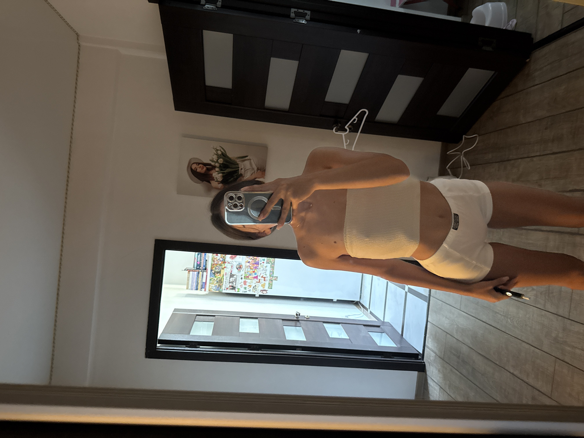 The image depicts a full-body mirror selfie of a young woman in an indoor setting. She is wearing a white strapless top and white shorts. The woman is holding a smartphone in her right hand, angled to capture the reflection. Behind her, a door with glass panels is visible, leading to a hallway with tiled flooring. Another door, darker in color and also with glass panels, is to her right. On the wall, a framed picture of a woman with a hat and flowers hangs. The floor is made of wooden planks. The environment appears to be a residential interior, possibly an apartment hallway or bedroom. No specific landmarks or elements definitively place the location within Chișinău, Moldova.