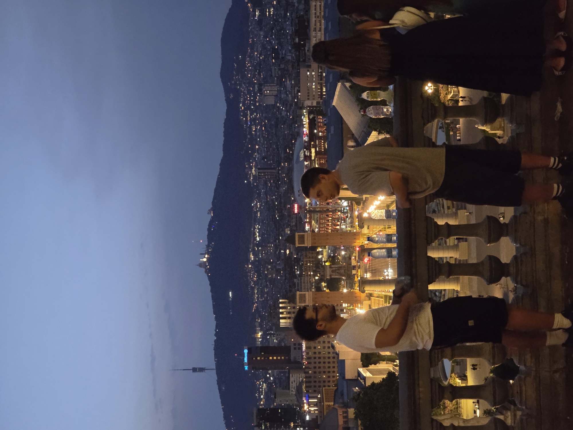 This is a nighttime photograph taken from a raised vantage point overlooking the city of Barcelona, Spain. In the foreground, three individuals are standing on a balcony or terrace, looking out at the cityscape. The individual on the left is a man wearing a white t-shirt and black shorts. The man in the middle is wearing a grey t-shirt and black shorts. The individual on the right is a woman wearing a long black dress. Two plastic water bottles are visible on the railing in front of the people.
The background features a sprawling urban landscape with numerous lights indicating buildings and streets. In the distance, a prominent hill is visible, topped with buildings that appear to be illuminated. A tall tower, likely a telecommunications tower, stands on the left side of the hill. The sky above is a deep blue, suggesting dusk or early night. The overall atmosphere is one of urban exploration and appreciation of the city view. The presence of the iconic Montjuïc hill and the general architecture visible strongly suggest the location is Montjuïc, with the view likely facing towards Plaça d'Espanya and the city center.