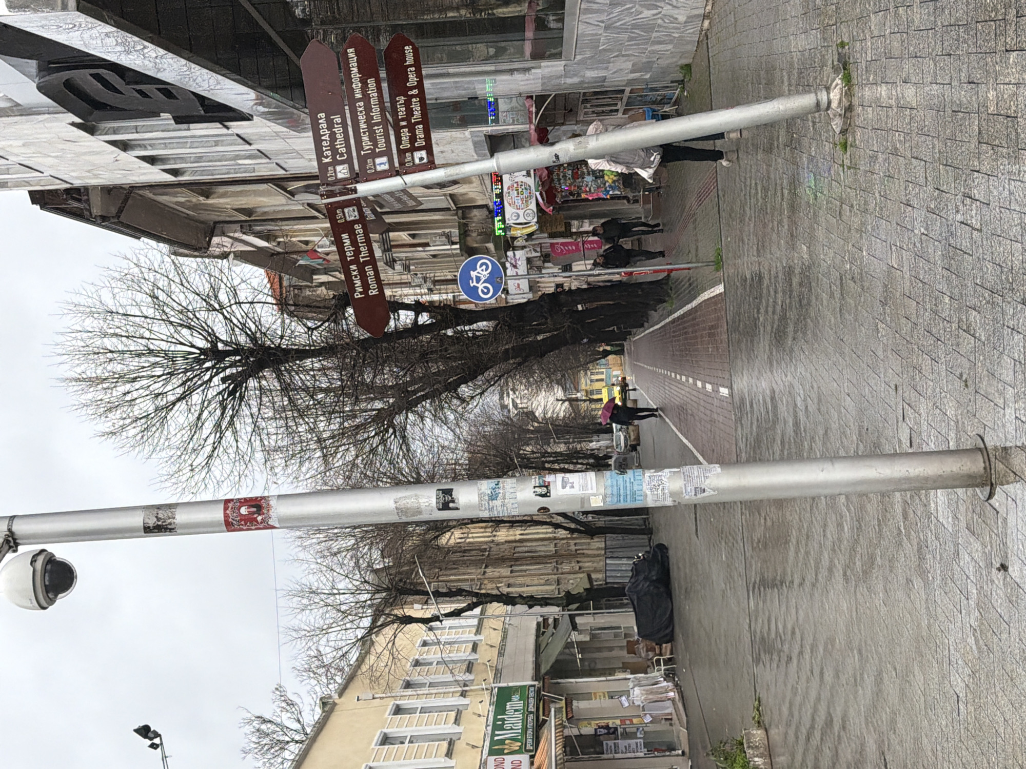 The image depicts a street in Varna, Bulgaria. There are buildings on both sides of the street, with leafless trees lining the sidewalk. A signpost indicates directions to local attractions, including the Cathedral (0.2km), Tourist Information (0.2km), the Drama Theatre & Opera house (0.1km), and the Roman Thermae (0.5km). People are walking on the sidewalk. A surveillance camera is mounted on a pole. There is a bicycle path marked on the road. Some shops and stores are along the street level. The weather appears to be overcast.