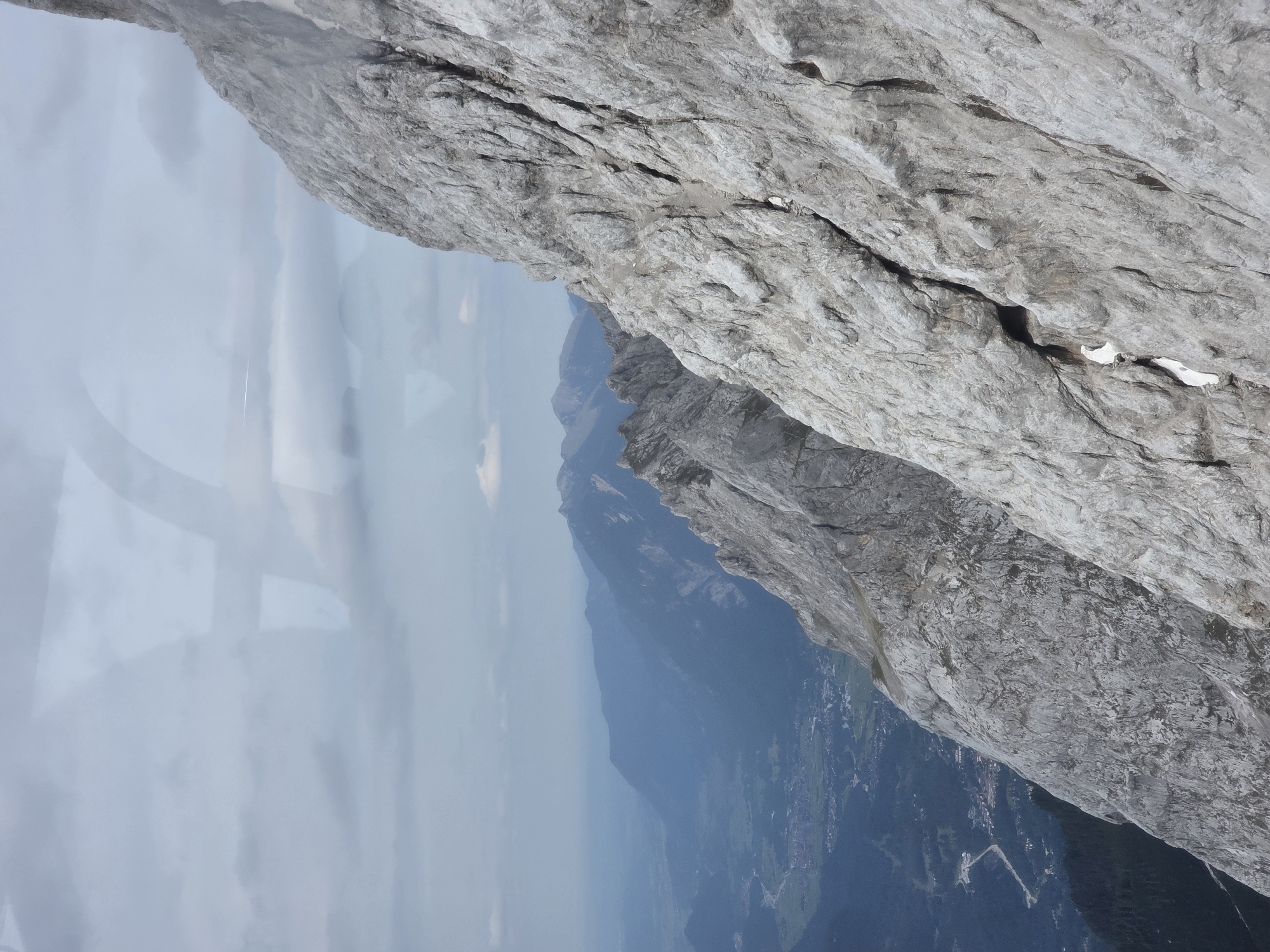The image captures a dramatic, close-up view of a rugged, rocky mountainside, likely taken from a high vantage point. The rock face dominates the foreground, characterized by its pale gray color, rough texture, and numerous fissures and crevices. Small patches of snow are visible nestled in some of the deeper cracks.
In the background, a vast landscape unfolds. Layers of progressively distant mountain ranges fade into a hazy blue, indicating a significant depth of field. The leftmost portion of the background reveals a valley with scattered settlements, roads, and green fields, suggesting a populated area nestled amongst the mountains. The sky above is overcast with diffused light, casting a muted tone over the entire scene. The overall impression is one of natural grandeur and the immense scale of the mountainous terrain.