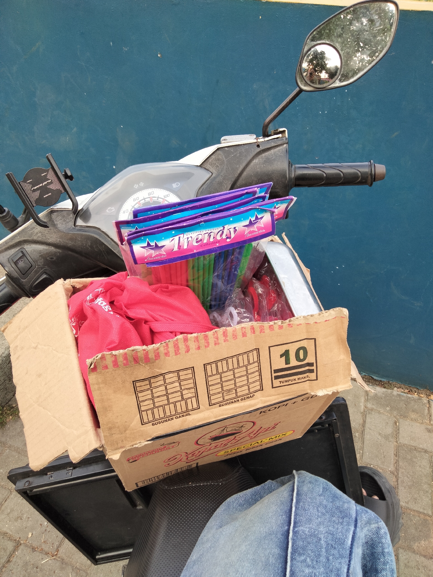 This is a medium shot, eye-level, outdoor photograph taken in Cariu, Indonesia. The photograph is focused on a scooter loaded with goods, with a blue wall in the background. The scooter's handlebar and instrument panel are visible in the upper left quadrant. A phone holder is attached to the handlebar. In the foreground, a brown cardboard box is placed on the scooter's rear rack. The box contains red fabric items and several packs of colorful straws. The straws are branded "Trends" and have a starry design. The box also displays markings indicating "10," "TUMPUK MAKS," and "SUSUNAN GENAP." To the right of the box, a metallic object is partially visible. The scooter's seat and a person's legs wearing blue jeans and black sandals are in the lower portion of the image. The ground is paved with light-colored bricks.