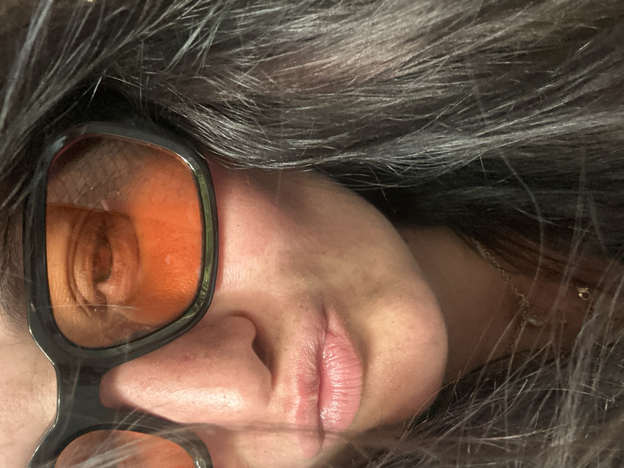 This is a close-up, waist-up, eye-level shot of a woman's face. She is wearing large, dark-rimmed glasses with orange lenses. Her mouth is pursed, and she appears to be looking down and slightly to her right, away from the camera. The woman has dark, long hair, which is partially obscuring her face. Her skin is fair, and she appears to be wearing light-colored lipstick. The background is dark and out of focus, with some light reflecting off her glasses. There is no indication of the city of Günzburg, Germany, or any specific environmental details.