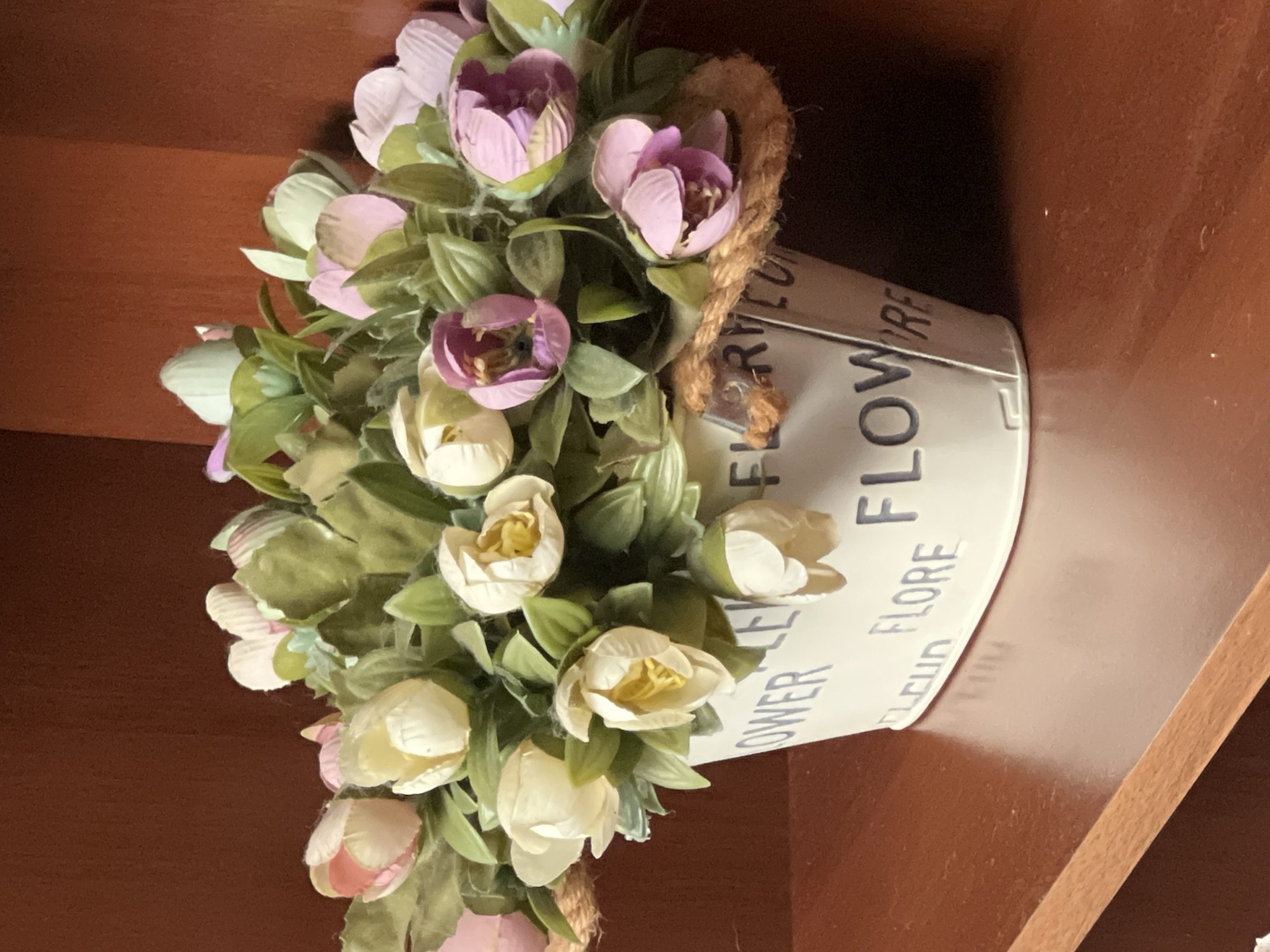 The image depicts a decorative metal pot filled with artificial flowers, primarily in shades of pink, white, and purple, with green foliage. The pot is white and has the words "FLOWER" and "FLORE" printed in blue on its side. A rope handle is visible on the side of the pot. The pot is placed on a polished wooden surface, which appears to be a shelf or table. Behind the pot, a dark red, textured surface is visible, possibly a wooden wall or cabinet. The lighting suggests an indoor environment. There is no indication of the city of Fontanka, Ukraine in the image.