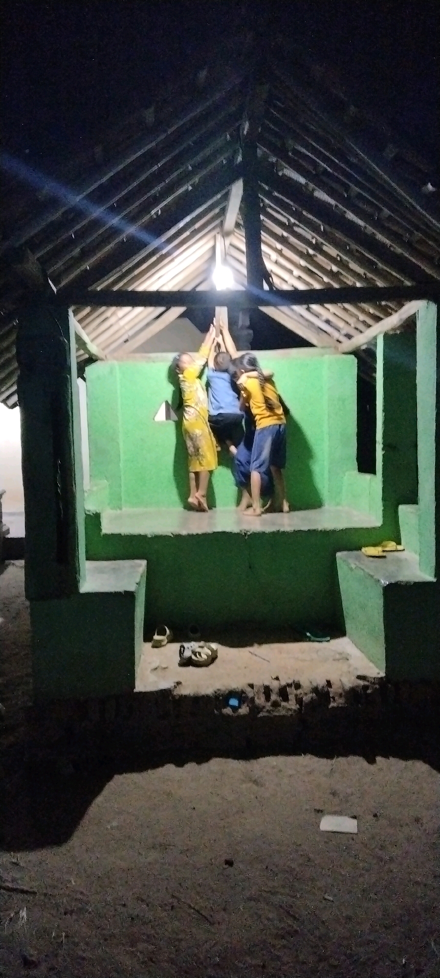 In the city of Tanjung Ratu Ilir, Indonesia, under a structure with a gabled roof supported by wooden beams, three young individuals, two appearing to be girls and one a boy, are gathered. The two girls, wearing yellow shirts and shorts, and the boy, in a blue shirt and dark shorts, are leaning against a bright green wall and reaching upwards, appearing to interact with a light fixture hanging from the ceiling. The structure has a raised platform painted green, with steps leading up to it. Outside the structure, on the ground, are a pair of white sandals, a pair of athletic shoes, and another pair of yellow sandals. The surrounding ground is dirt and appears to be an outdoor area at night, illuminated by the light within the structure.