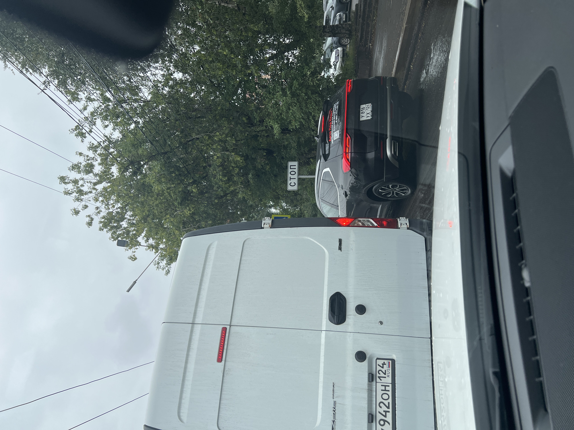 A white van is parked behind a black SUV on a street in Krasnoyarsk, Russia. The scene is viewed from inside another vehicle, with the dashboard on the right side of the frame. The weather appears overcast, and the ground is wet, suggesting it has recently rained or is raining. Trees with green foliage line the street on the left, and power lines are visible overhead.
A "STOP" sign is prominently displayed on a pole next to the black SUV, indicating an intersection or a designated stopping point. The license plate of the black SUV is visible and reads "Y 816 YX 124 RUS". The license plate of the white van is also visible, showing "Y942OH 124 RUS". The van has a large, flat rear door with a handle. The SUV has a distinctive taillight design and a chrome trim along its windows. The overall impression is of traffic congestion on a cloudy day.