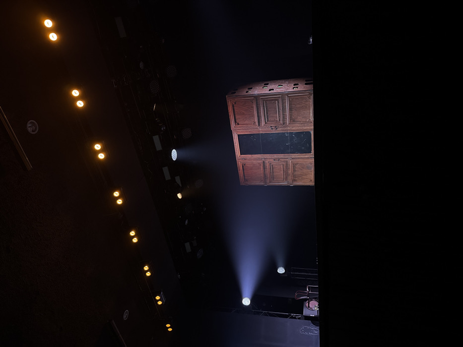 The image captures a dramatic, dimly lit scene, strongly suggesting a stage or performance venue. The perspective is looking upwards, towards the ceiling or overhead rigging.
On the left side of the frame, a series of warm, yellowish lights are visible, arranged in a diagonal line, illuminating a dark, textured surface, possibly part of the venue's ceiling or wall. These lights appear to be smaller, accent or house lights.
Towards the center and right, the scene becomes more focused on stage lighting. Several powerful, cool-toned spotlights emit bright white and bluish beams of light that cut through the dark space. The visible shafts of light indicate the presence of haze or smoke in the air, a common atmospheric effect in live performances.
Dominating the upper right portion of the image is a large, ornate wooden structure suspended from above. It resembles a detailed cabinet or an intricate wall panel, featuring multiple rectangular sections. The wooden elements are a medium brown color with visible grain, and some sections have decorative molding. In the center of this structure, there appears to be a darker, rectangular panel, possibly a screen or a reflective surface, which adds to its theatrical appearance.
The overall atmosphere is one of anticipation or active performance, with the dramatic lighting drawing attention to the suspended structure and the invisible space below the light beams, where a stage or performers would likely be located. The background is predominantly dark, emphasizing the illuminated elements. No text is visible in the image.