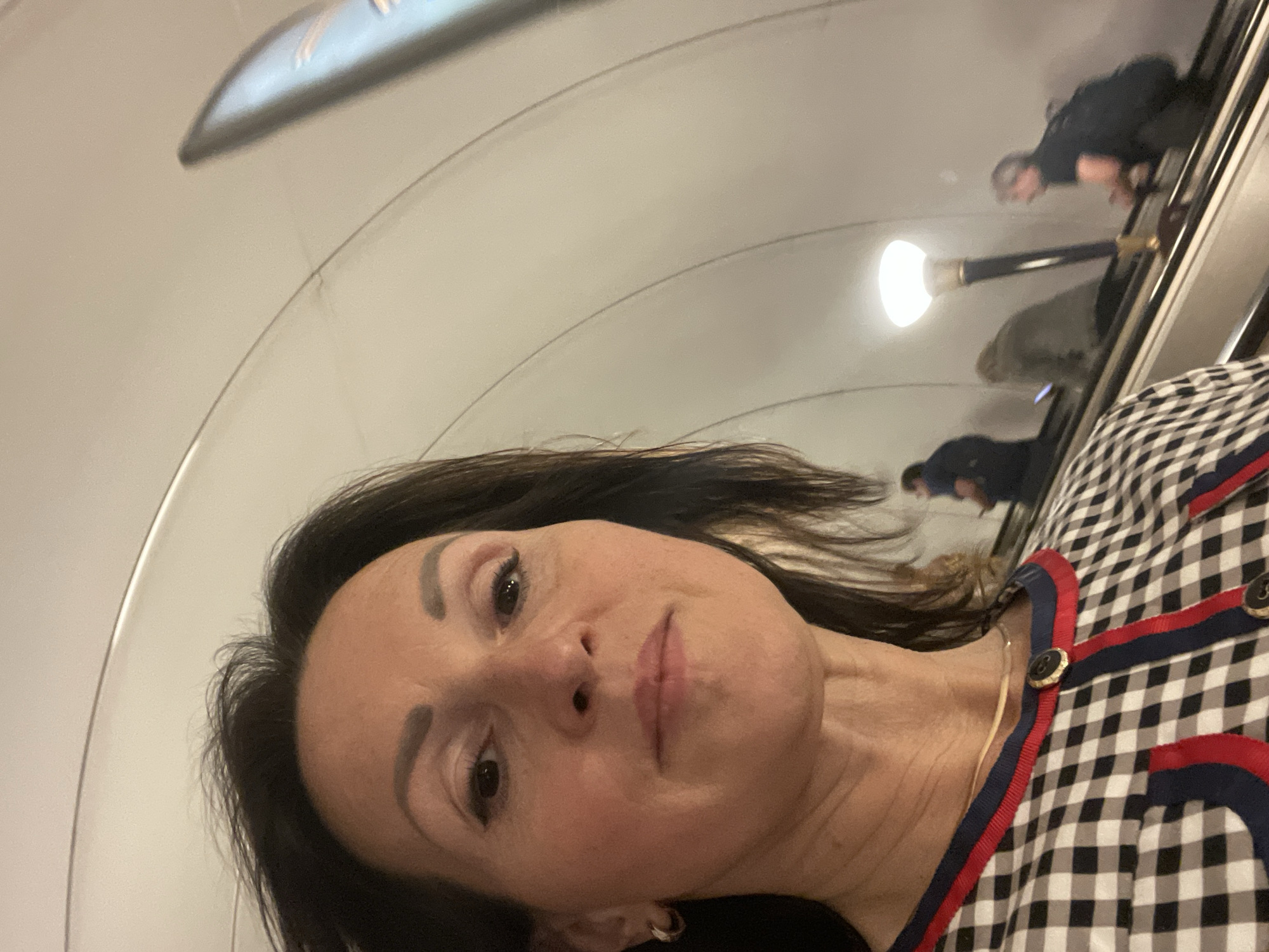 A medium shot captures a woman taking a selfie while standing on an escalator. She is looking directly at the camera with a neutral expression. The woman has dark brown hair, styled down, and she is wearing a black and white checkered top with red and navy blue trim. She has on a thin gold necklace and a small hoop earring in her left ear. In the background, the curved, cream-colored ceiling of a subway station is visible, with some ambient lighting. Several other people are on the escalator behind her, some blurred due to motion. The overall environment suggests an indoor public transportation hub.
