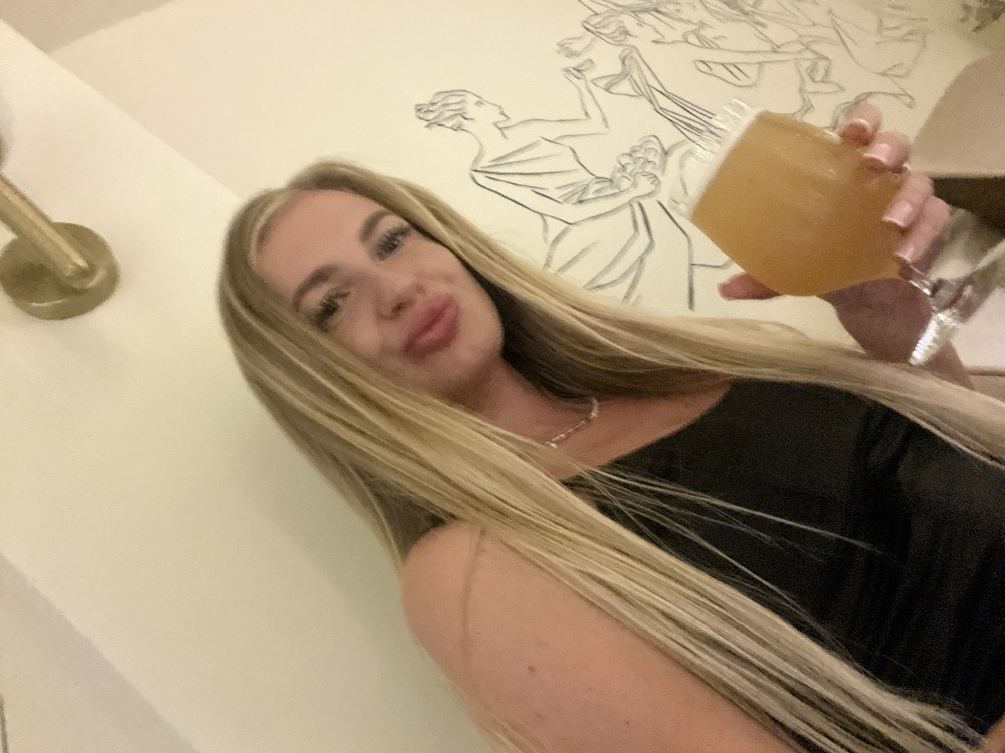 A woman is holding a glass of amber liquid in a room. The location is Boreti, Montenegro. The woman has long, straight blonde hair, light skin, and is wearing a black top. She is looking at the camera with her lips pursed. Her right hand is holding a clear glass filled with a light brown beverage. The background is a white wall. On the wall is a drawing of Greek figures. A gold-colored light fixture is on the wall to the left.