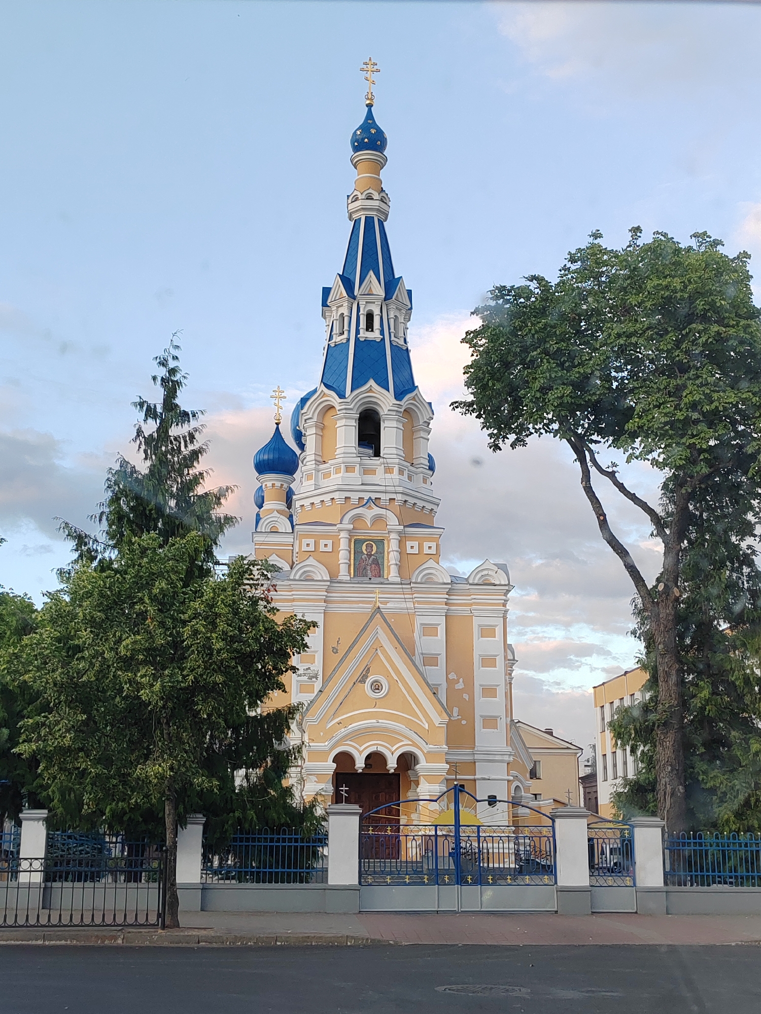 The image shows the Holy Cross Cathedral in Brest, Belarus. The cathedral is a yellow building with blue domes and a tall bell tower topped with a golden cross. In front of the cathedral is a blue metal fence with decorative elements that include crosses and a sun. Two trees, one tall and slender, the other large and leafy, frame the cathedral on either side. The sky is cloudy with hints of pink and orange from the setting sun. The foreground shows a paved street and sidewalk. No people are visible in the image.