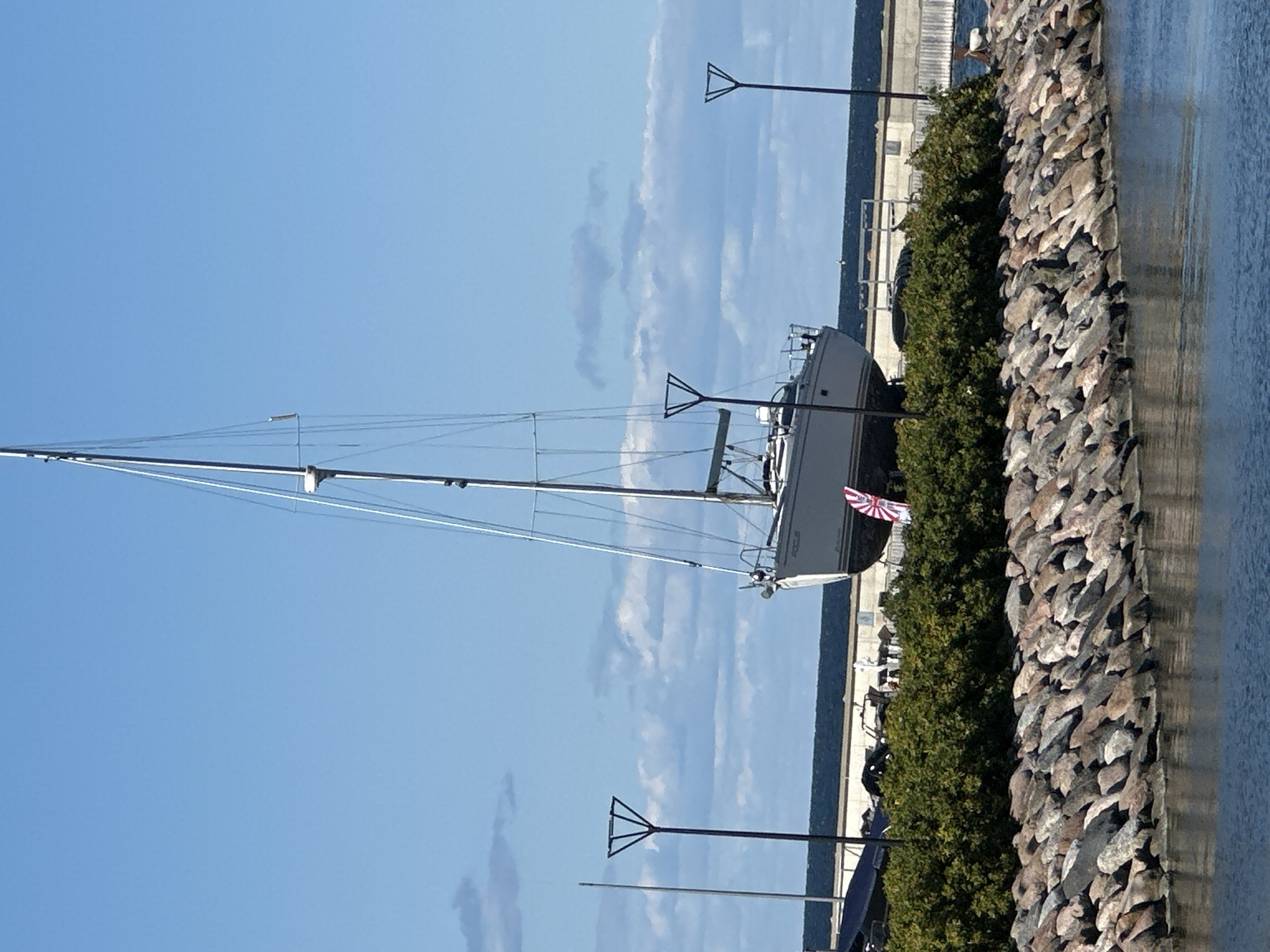 The image shows a sailboat named "FOCUS" resting on land, possibly undergoing maintenance or storage, near a body of water. The sailboat is white with a visible name on its hull. It has a tall mast with rigging. In the background, there is a concrete structure, possibly a pier or seawall, and a building. The foreground features a rocky embankment leading to the water, with some green bushes on top of the embankment. Two distinct modern lampposts with triangular tops are visible on the land. The sky is blue with scattered clouds. The scene appears to be a harbor or marina area. The location is specified as Tallinn, Estonia. There are no people visible in the image.