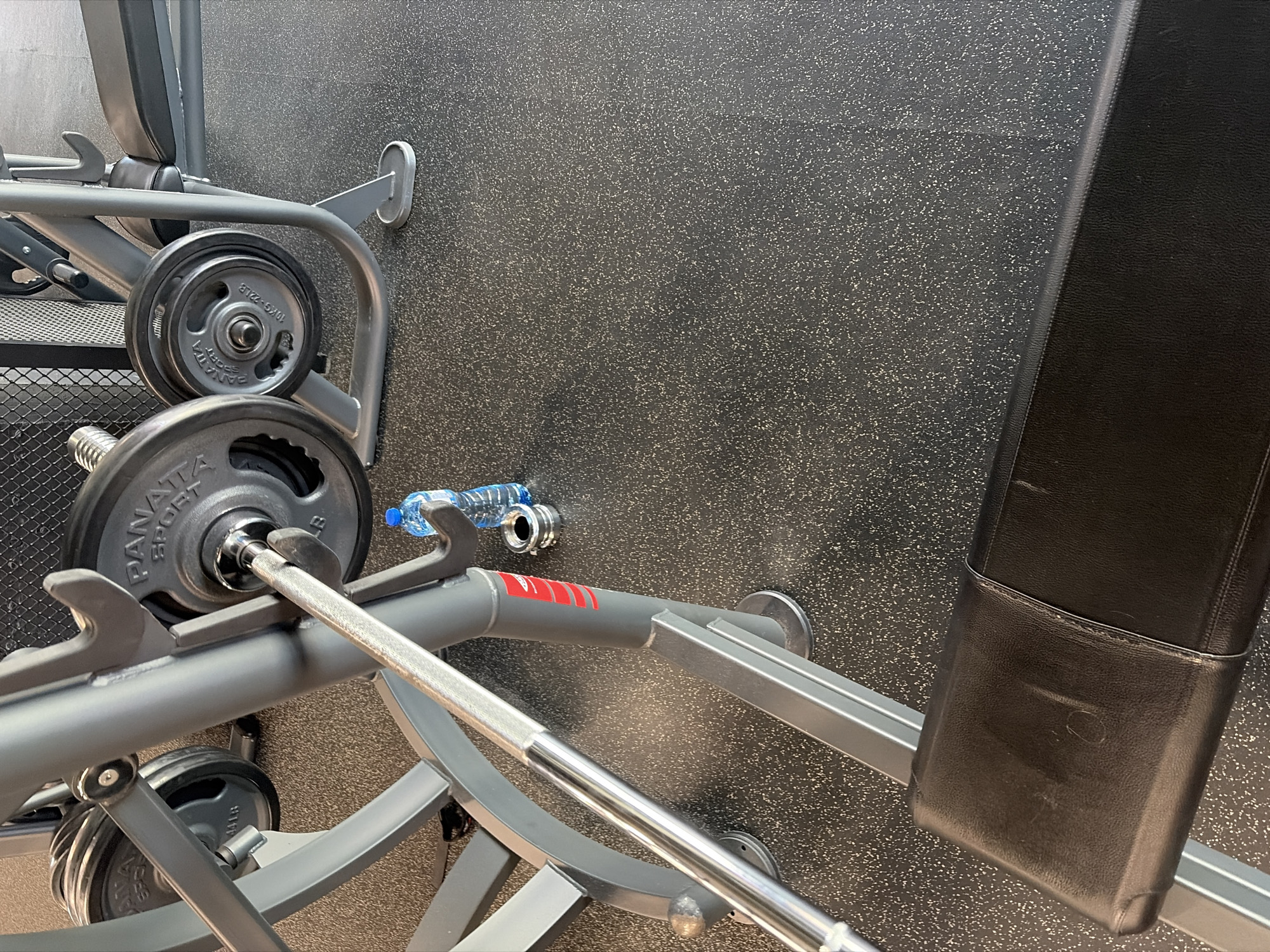This image depicts an indoor scene within a gym or fitness center, located in Plovdiv, Bulgaria. The shot is taken from a slightly elevated, downward-angled perspective, focusing on weightlifting equipment and the floor.
The primary subjects are a gray metal squat rack or bench press machine, a barbell, and several weight plates. The barbell, made of shiny silver metal, is resting on the safety catches of the machine. It is loaded with dark gray, circular weight plates. Visible text on one of these plates clearly reads "PANATTA SPORT" and "10KG 22LB". Another plate below it also shows "10KG". Additional weight plates are stored on a vertical peg attached to the machine's frame. The machine itself features robust gray metal tubing and safety mechanisms.
To the right, part of a black, padded bench is visible, likely integral to the weightlifting machine. The floor is a dark gray or black speckled rubber material, commonly found in gyms for durability and cushioning. A small, clear plastic water bottle with a blue cap is lying on its side on the floor, near the base of the equipment.
No people are present in the image, and no specific activity is actively occurring, though the equipment is set up for use. The scene suggests a ready-to-use weight training area. There are no indications of the time of day or external weather conditions.