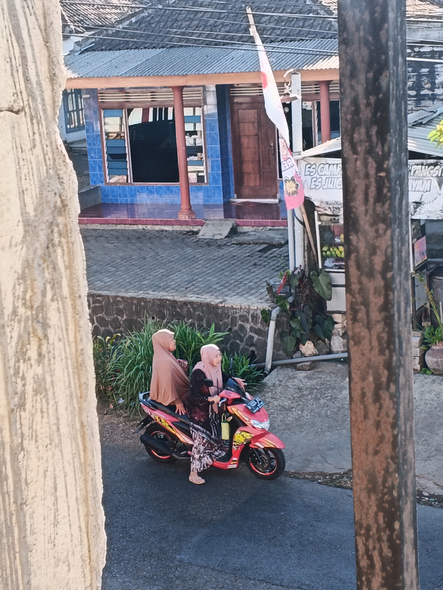 In Bocek, Indonesia, two women are seated on a red and yellow scooter parked on a paved road. The woman in front, wearing a tan hijab and a patterned dress, is looking to her right. The woman behind her, wearing a pink hijab and a darker patterned dress, is looking to her left. A yellow bag hangs from the scooter's handlebars. The background shows a building with blue tiled walls and a corrugated metal roof, with a wooden door and windows. A flag is attached to a pole in front of the building. There is also vegetation and a stone wall to the left of the scooter. A utility pole stands to the right of the scene, partially obscuring the background.
