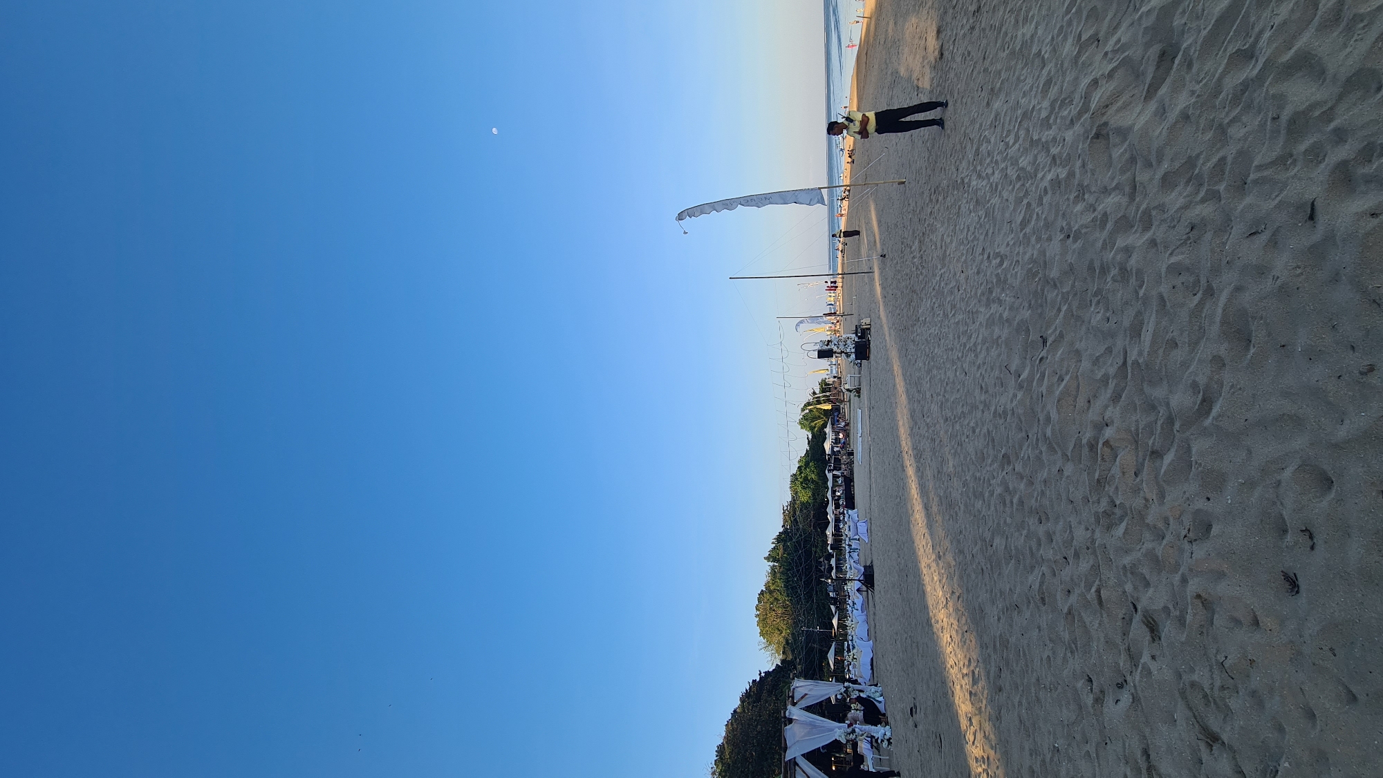 This media file captures a panoramic view of a beachside event set up for what appears to be a reception or celebration. The scene is dominated by a clear, bright blue sky with a crescent moon visible in the upper left quadrant. The foreground is composed of a sandy beach, with a long shadow cast across it from the right, suggesting it is late afternoon or early morning.
In the mid-ground, a man wearing a yellow shirt and dark pants stands on the beach, facing away from the camera and towards the ocean. Further down the beach, a series of white tents and draped fabric structures are erected, suggesting a catered event. Tables and chairs are set up beneath some of these structures, adorned with white tablecloths and floral arrangements, indicating guests are either present or expected. White flags or banners are also visible, adding to the festive atmosphere. The background shows the ocean with a faint horizon and a few scattered structures or buildings along the coastline. Lush green trees line the edge of the beach area, providing a natural backdrop to the event setup.
The overall impression is one of a well-organized outdoor event taking place in a tropical or coastal setting, likely in Denpasar, Indonesia, given the context. The clear sky and gentle lighting suggest pleasant weather. The event appears to be a formal gathering, possibly a wedding or a corporate function, given the decor and setup. No visible text is present in the image.