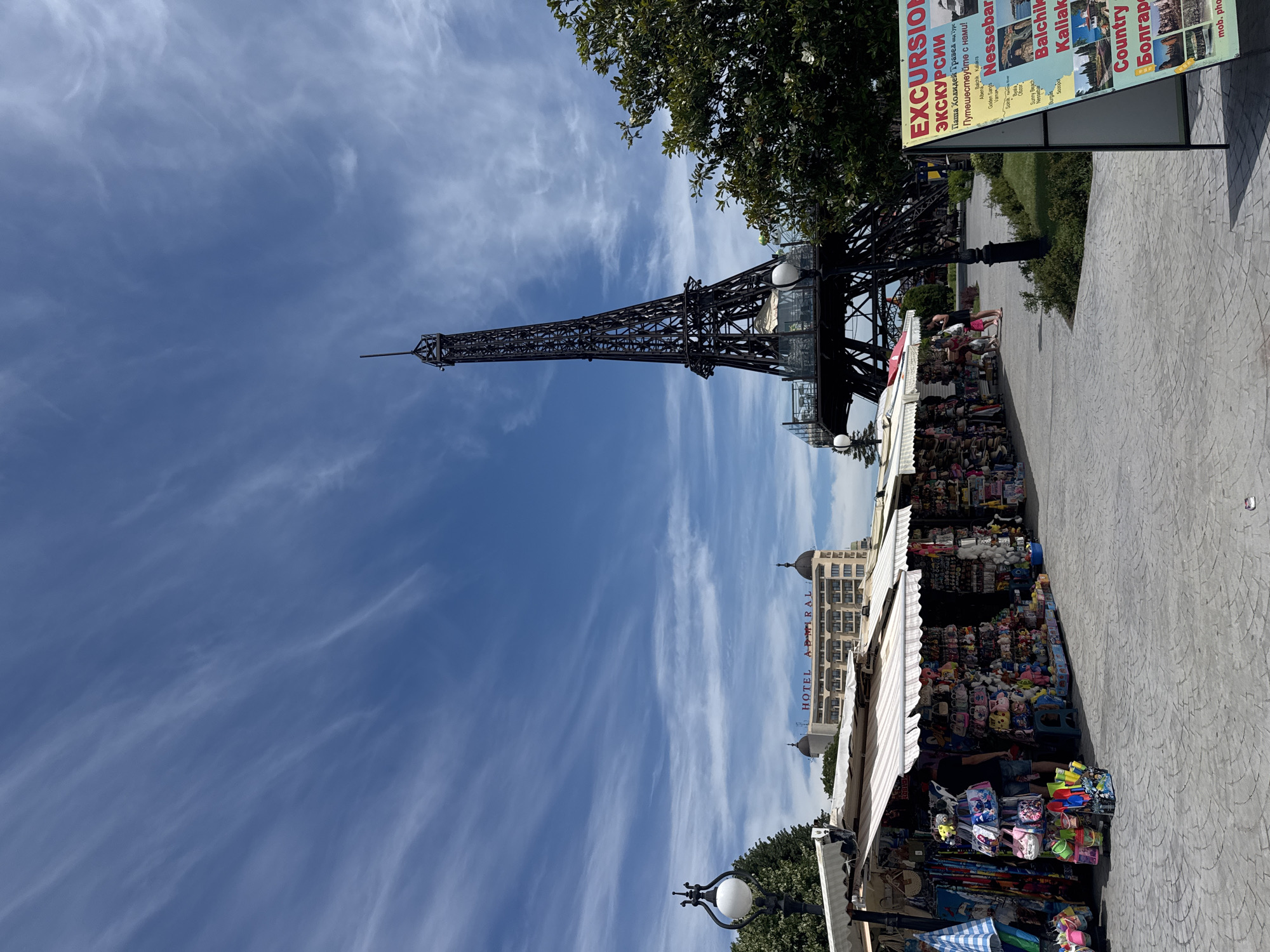 In Varna, Bulgaria, a replica of the Eiffel Tower stands tall against a bright blue sky dotted with wispy clouds. In the foreground, several market stalls are set up, displaying a variety of souvenirs, clothing, and beach toys. A person is seen browsing the items at one of the stalls. In the background, a hotel named "HOTEL ADMIRAL" is visible, and lush green trees provide a natural backdrop. The scene is set on a paved walkway, indicating a public area, likely a promenade or tourist attraction. A sign advertising "EXCURSIONS" is prominently displayed near the market stalls, suggesting this is a popular tourist destination.