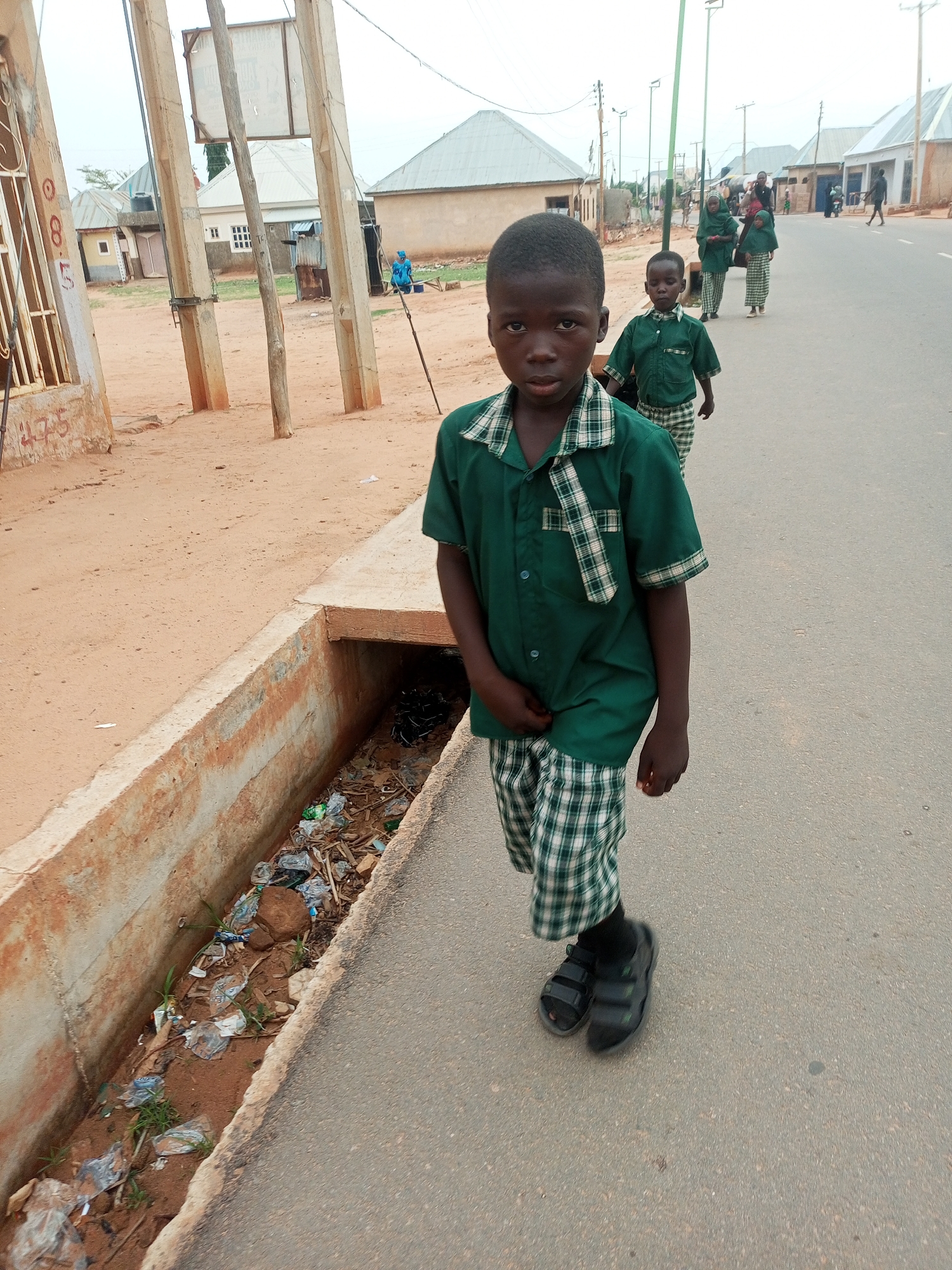 A young boy in a green and white plaid uniform and black sandals stands on a paved road next to a concrete drainage ditch filled with trash. Behind him, other children in similar uniforms walk in the same direction. The location is Gombe, Nigeria, evidenced by the style of buildings and infrastructure in the background. Buildings in the background are single story and made of concrete. Overhead power lines are present. The sky is overcast.