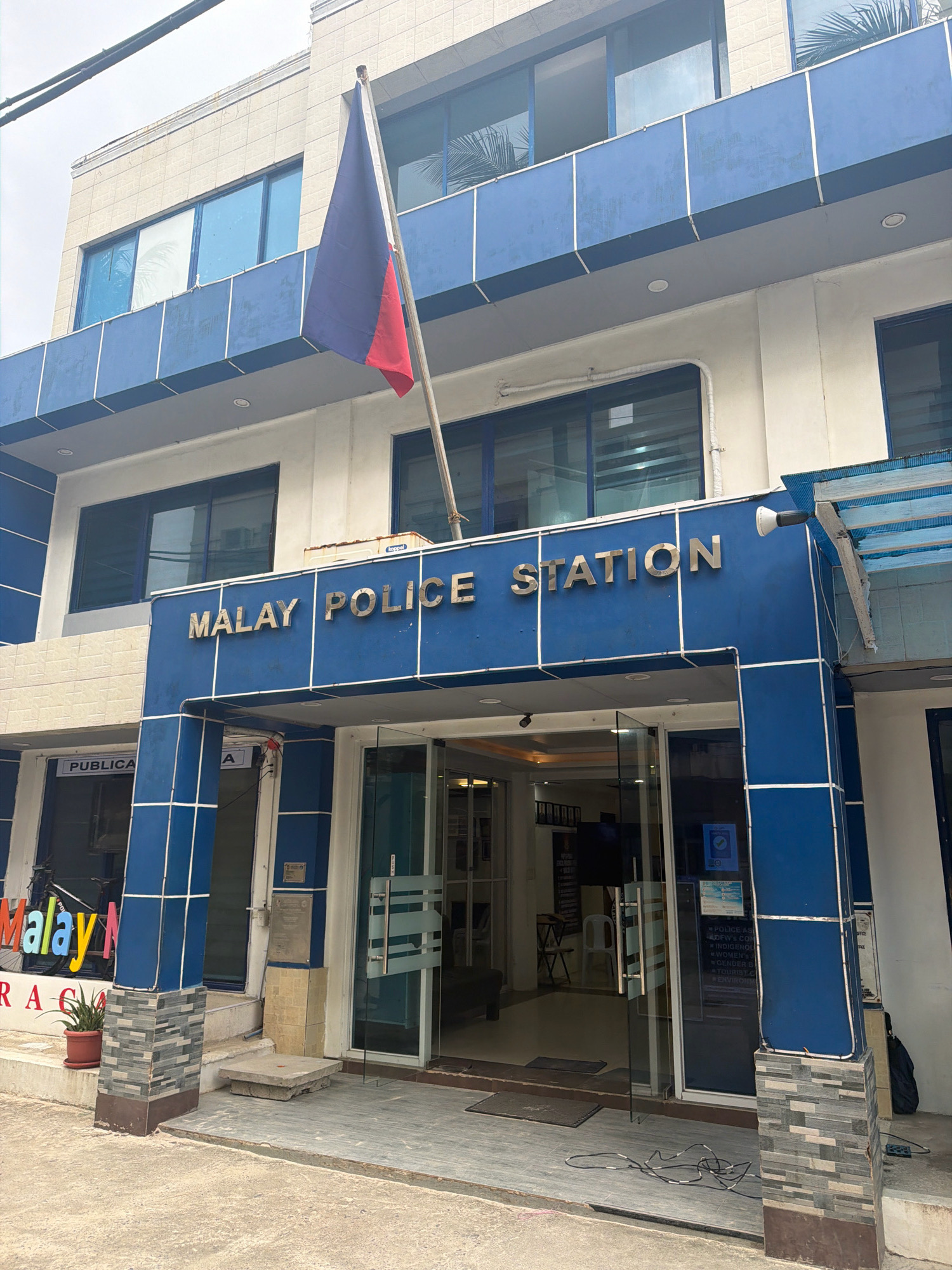 The image shows the exterior of the Malay Police Station in Malay, Philippines. The building is a two-story structure with a blue and white facade. The main entrance features large glass doors and is adorned with the words "MALAY POLICE STATION" in large gold lettering. A Philippine flag is visible flying from a flagpole to the left of the entrance. The area in front of the building is paved, with some planters and a small concrete platform. Other signs and details on the building suggest it houses various services related to law enforcement and public safety.