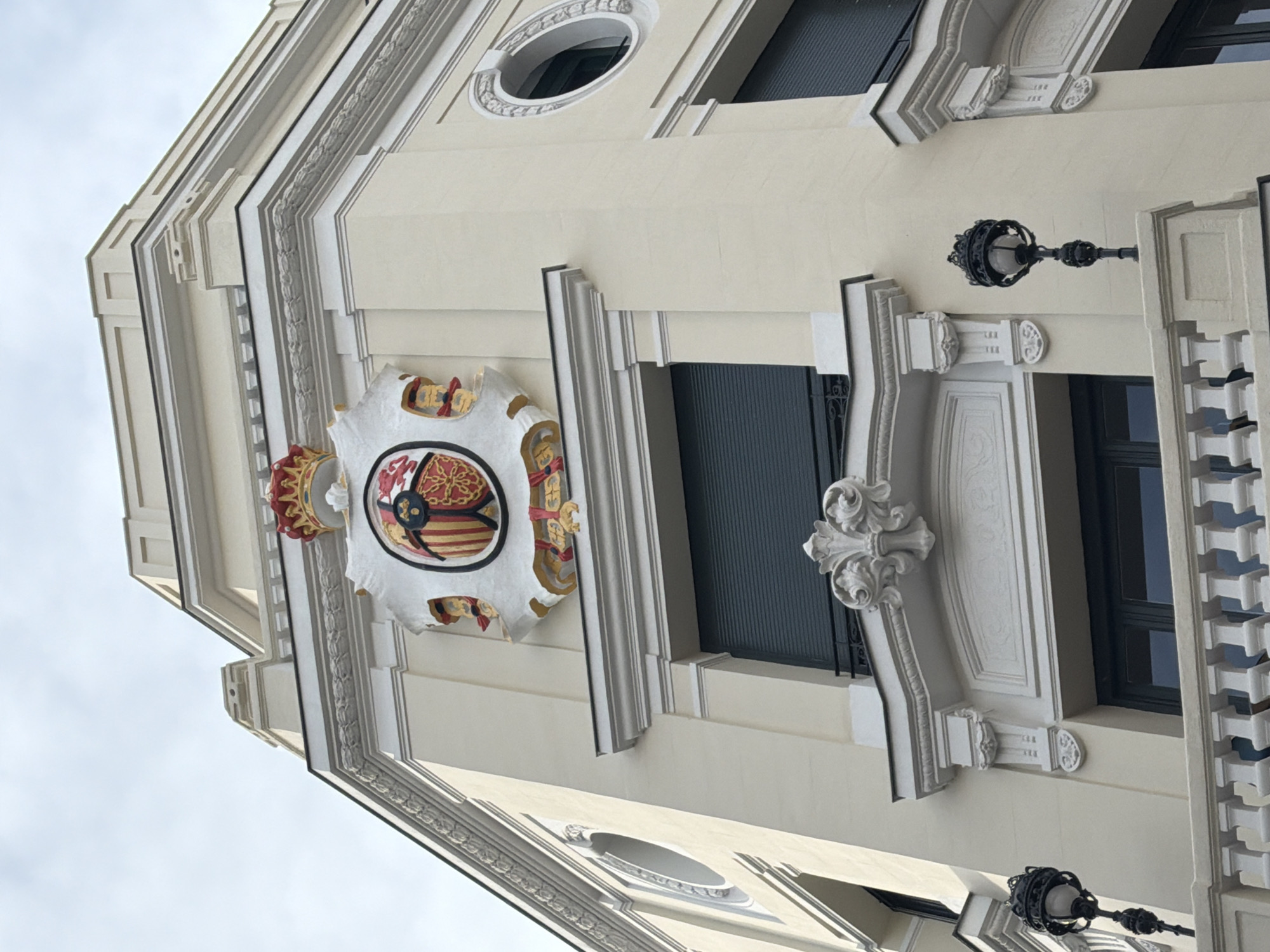 The image depicts a section of a building facade in Madrid, Spain. The architectural style is ornate, featuring detailed trim, moldings, and decorative elements. A prominent heraldic shield with a crown is affixed to the building's surface. Windows are interspersed along the facade, some covered by closed blinds. Ornate lampposts are attached to the building at intervals. The sky is visible in the background.