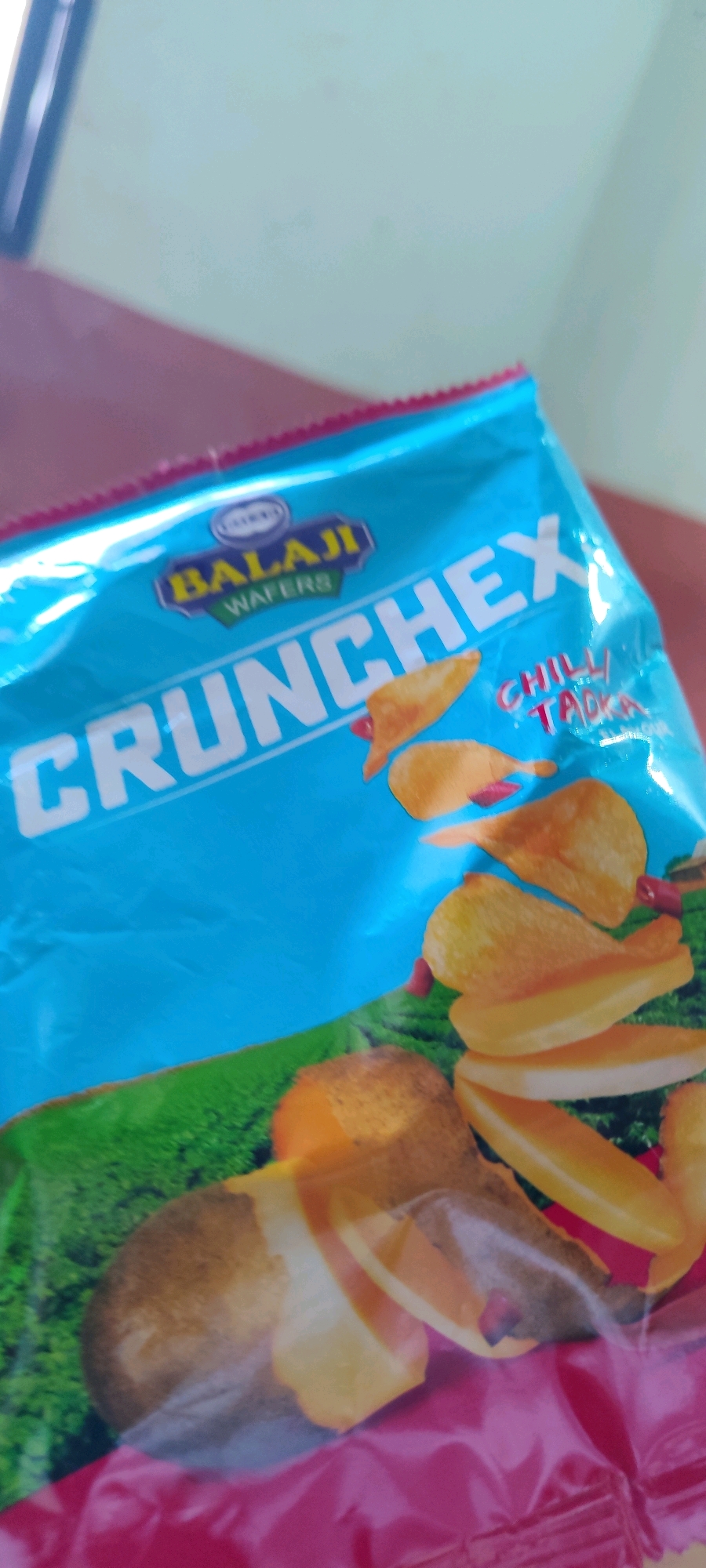 The image is a close-up, slightly blurry shot of a bag of Balaji Wafers CrunchEx, specifically the "Chilli Tadka" flavor. The bag is predominantly bright blue with white lettering for the brand name and product. The foreground shows some potato chips, some appearing fried and sliced. The background is out of focus, showing what appears to be a surface and a wall, suggesting an indoor setting. The image is taken in Belagavi, India.
