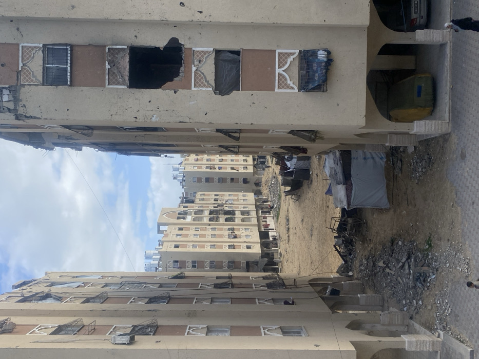 The image depicts a devastated urban residential area in Khan Yunis, Palestinian Territory. Two multi-story apartment buildings with light-colored facades and darker brown vertical accents frame a central expanse of unpaved earth filled with debris. Both foreground buildings exhibit extensive damage, including shattered windows, twisted shutters, and large sections of external wall missing, revealing darkened interiors. The building on the right has a prominent, large impact hole on its upper levels.
In the middle ground, several makeshift shelters constructed from tarpaulins and various materials are visible on the sandy ground, some with laundry hanging. Piles of rubble, twisted metal, and discarded objects are scattered across this central area. In the background, additional multi-story residential buildings, some also showing signs of damage, line the horizon under a partly cloudy sky. Water tanks are visible on the rooftops of these distant structures.
On the paved foreground, a person wearing a hijab walks on the right, and a child-sized figure walks on the left. Under an archway of the right foreground building, a large, yellow-green object covered by a tarp is situated, alongside a parked car. Several small figures are discernible in the distance within the central debris field, indicating human presence.