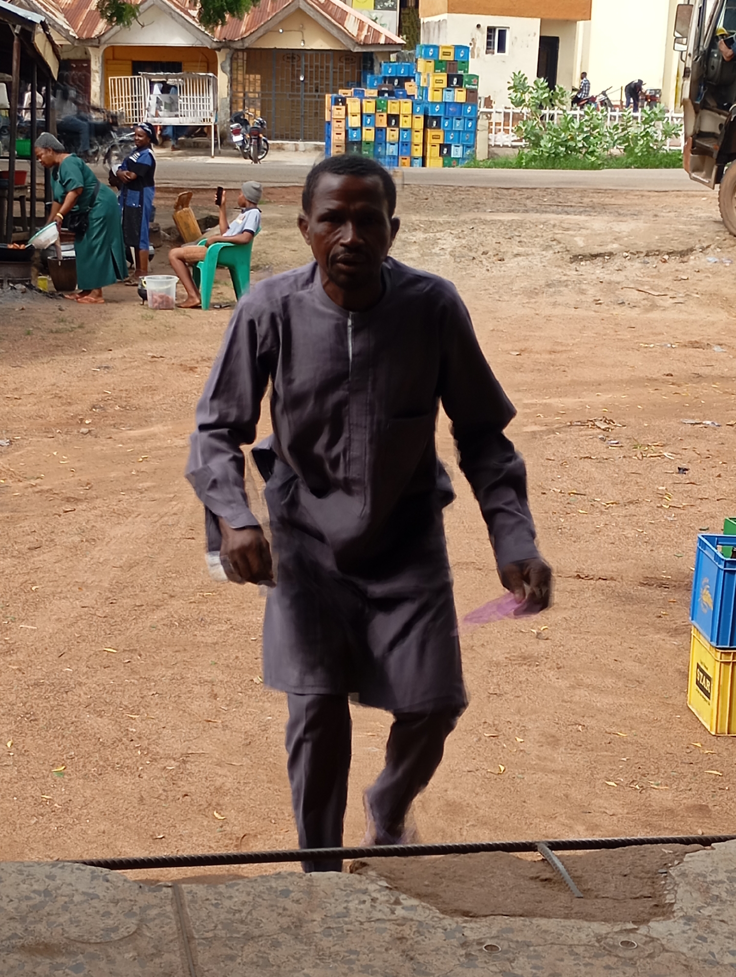 In Shendam, Nigeria, a man in a dark gray traditional outfit is walking forward, his left hand extended holding a small purple object. He has a focused expression. To his left, a woman in a green dress is cooking at an outdoor stall, and another woman in a blue and black outfit stands nearby. Behind them, a man in a light blue shirt and dark pants sits in a green plastic chair, holding a mobile phone. In the background, buildings with corrugated metal roofs line a street, and stacks of colorful crates are visible. The ground is covered in reddish-brown dirt, with scattered leaves and debris. A motorcycle is parked to the left of the buildings. A truck is partially visible on the far right of the frame.