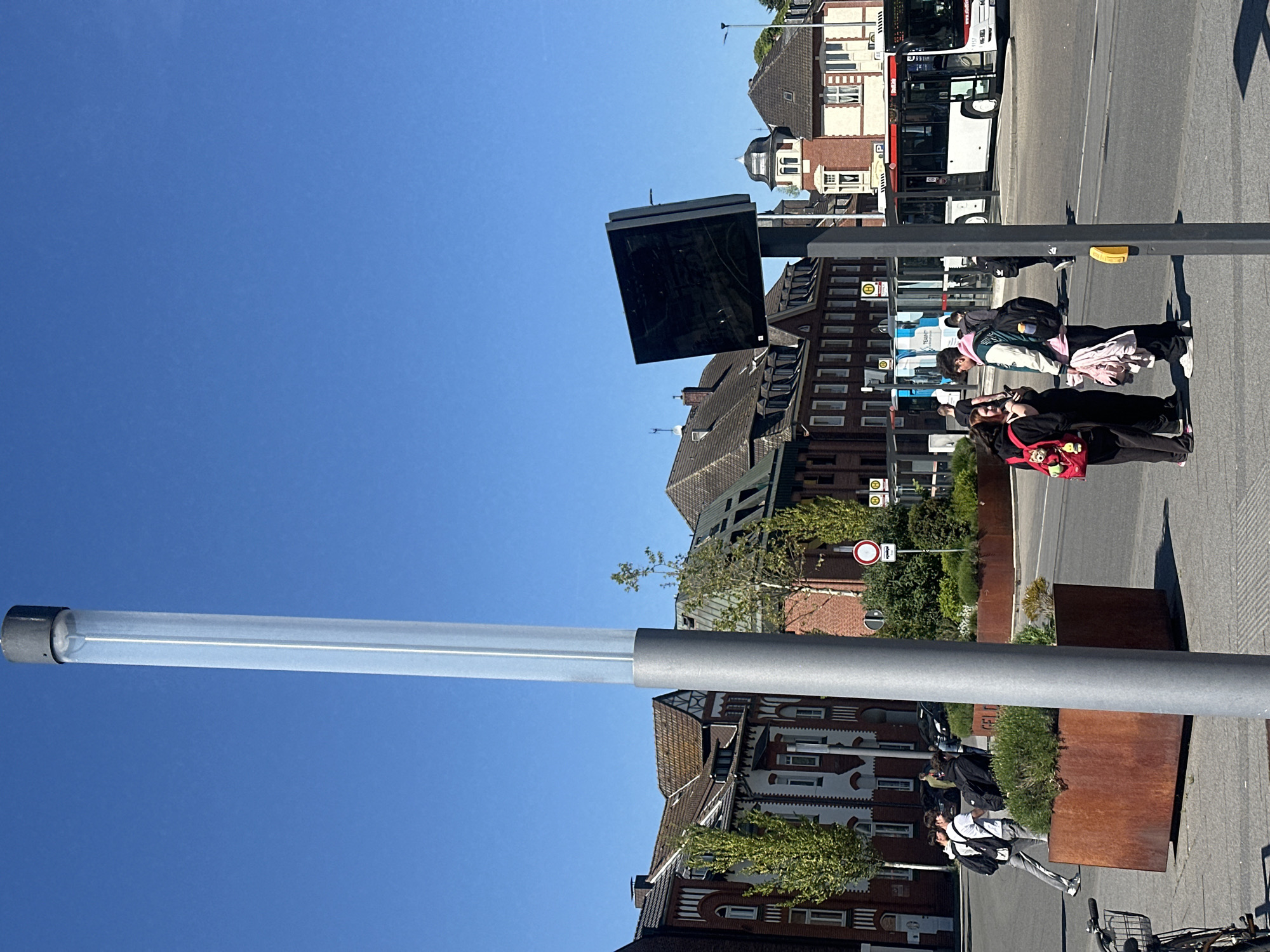 This image captures a vibrant urban street scene in Geldern, Germany, on a clear, sunny day. The sky is a bright, cloudless blue, and strong shadows indicate a high sun.
In the foreground, on the left side of the frame, stands a tall, modern street light or public art installation, featuring a clear, cylindrical tube atop a gray metallic pole. To its right, a black digital display screen, possibly for public transport information, is mounted on another pole.
The mid-ground features a group of approximately three to four people gathered on a paved sidewalk, appearing to be at a bus stop. One individual wears a light-colored jacket with pink and white sections, while another carries a distinctive red bag adorned with a teddy bear or character. Behind them, a blue and white bus is partially visible. Further down the street, two more individuals are walking on the sidewalk, one in a white shirt and backpack.
The background is dominated by a mix of traditional and modern multi-story buildings. Some feature pitched roofs, brick facades, and white window frames, characteristic of German architecture. A white bus with "112" visible on its side is parked or driving on the street in front of these buildings.
Various urban elements are present, including large rectangular planters made of rusty brown metal, filled with green bushes. One planter visibly shows the text "GELD" (likely part of "Geldern" or a related word). Traffic signs include a circular red and white "no entry" sign and multiple yellow "H" signs, indicating bus stops (Haltestelle). A yellow pedestrian signal button is also visible on a pole. A bicycle is partially seen parked at the bottom right of the frame.