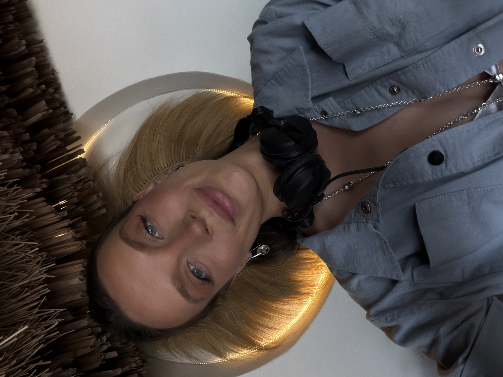 A woman with blonde hair, wearing a grey shirt and headphones, poses for a selfie. She has on a necklace and earrings. The background appears to be an indoor setting with a light fixture and straw-like decor. There is no indication of the city of Drobnići, Montenegro.