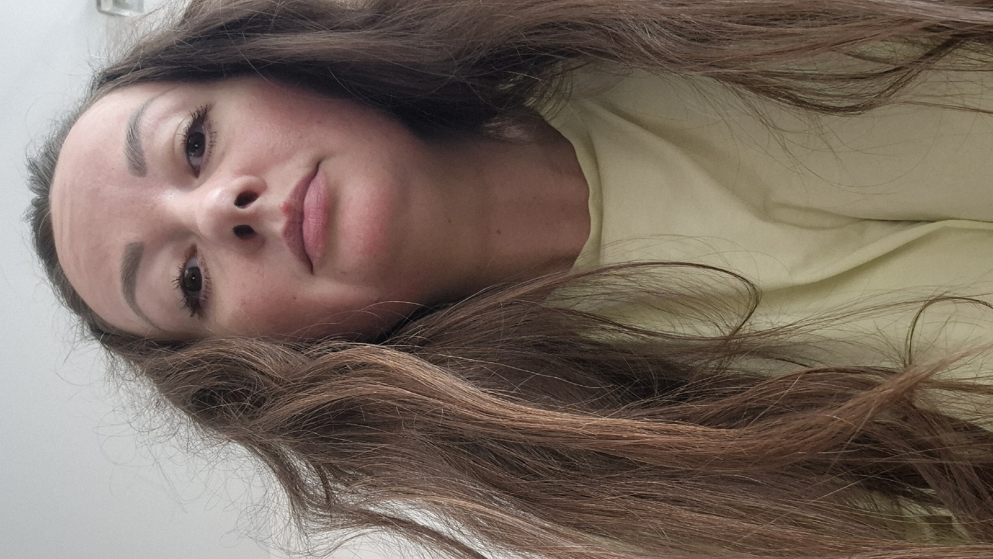 The image captures a young woman lying down and looking directly at the camera. Her head is positioned on the left side of the frame, with her long, wavy, dark brown hair spread out across the lower and right portions of the image. She is wearing a light yellow garment, possibly a t-shirt, visible around her neck and shoulder.
Her facial expression is neutral, with a slight hint of a smirk. She has prominent dark eyebrows and long, dark eyelashes. Her lips are a soft pink color, and a small reddish mark is visible on her upper lip. Her cheeks show a faint flush.
The background behind her head is a plain, light-colored surface, potentially a wall. The remainder of the background consists of the light yellow fabric she is resting on, along with her voluminous hair. The setting appears to be indoors, suggesting a casual environment like a home. The lighting is soft and even, creating a gentle ambiance. There are no specific visual cues within the image to confirm the provided location context of Kemerovo, Russia. No text is visible in the image.