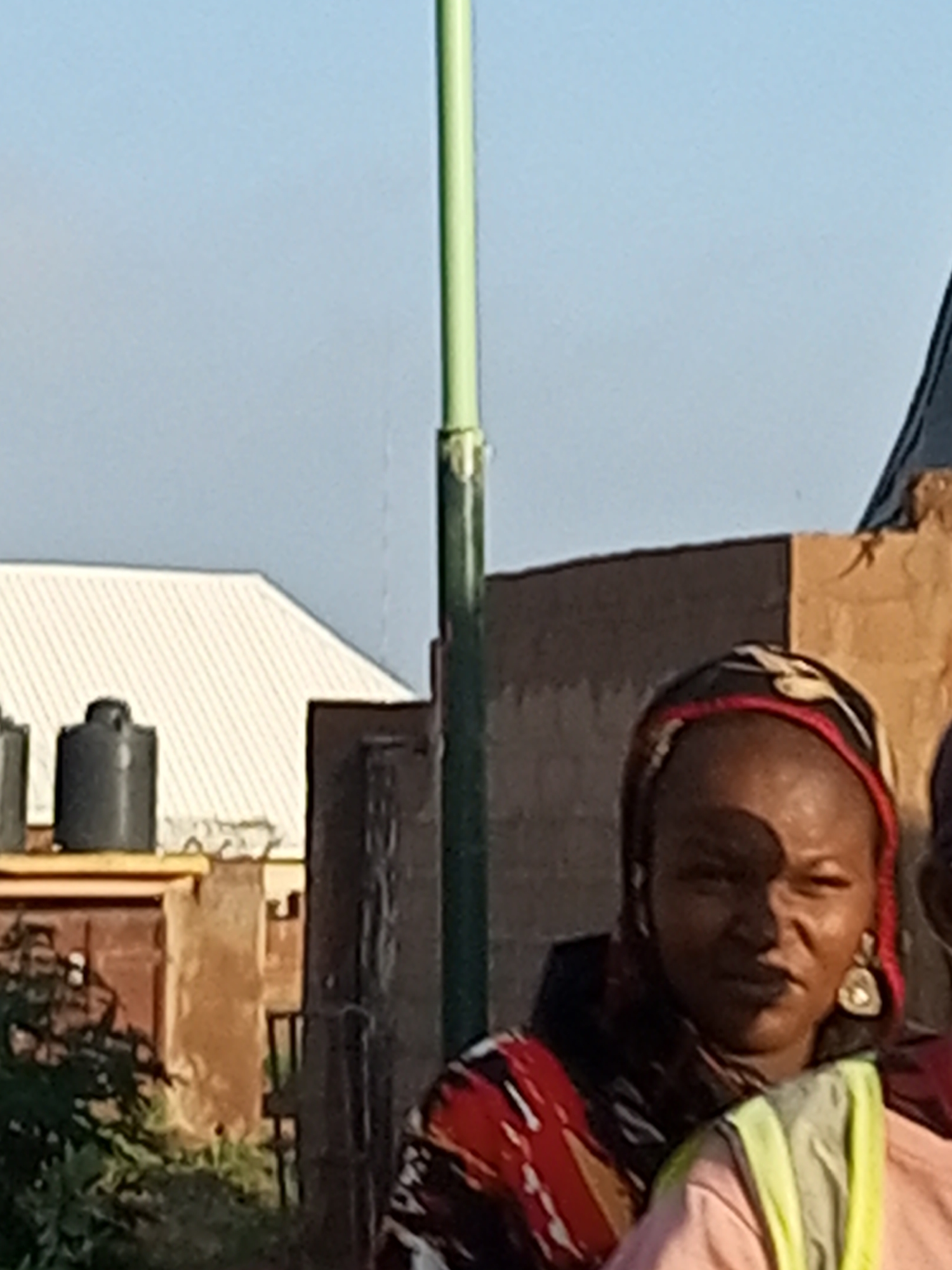 An adult female, wearing a red and black headscarf with a white pattern, a red, black, yellow, and white patterned garment, and a light pink shirt, stands in front of a partially constructed building. She is wearing a large, tear-drop shaped earring in her right ear. She is looking towards the right of the frame. Behind her, a light green pole, likely a light pole, stands tall. In the background, to the left, a white, corrugated metal roof is visible on a building. Two black water tanks are also present. The environment appears to be outdoors, with a clear sky. The scene is likely located in Tempure, Nigeria.