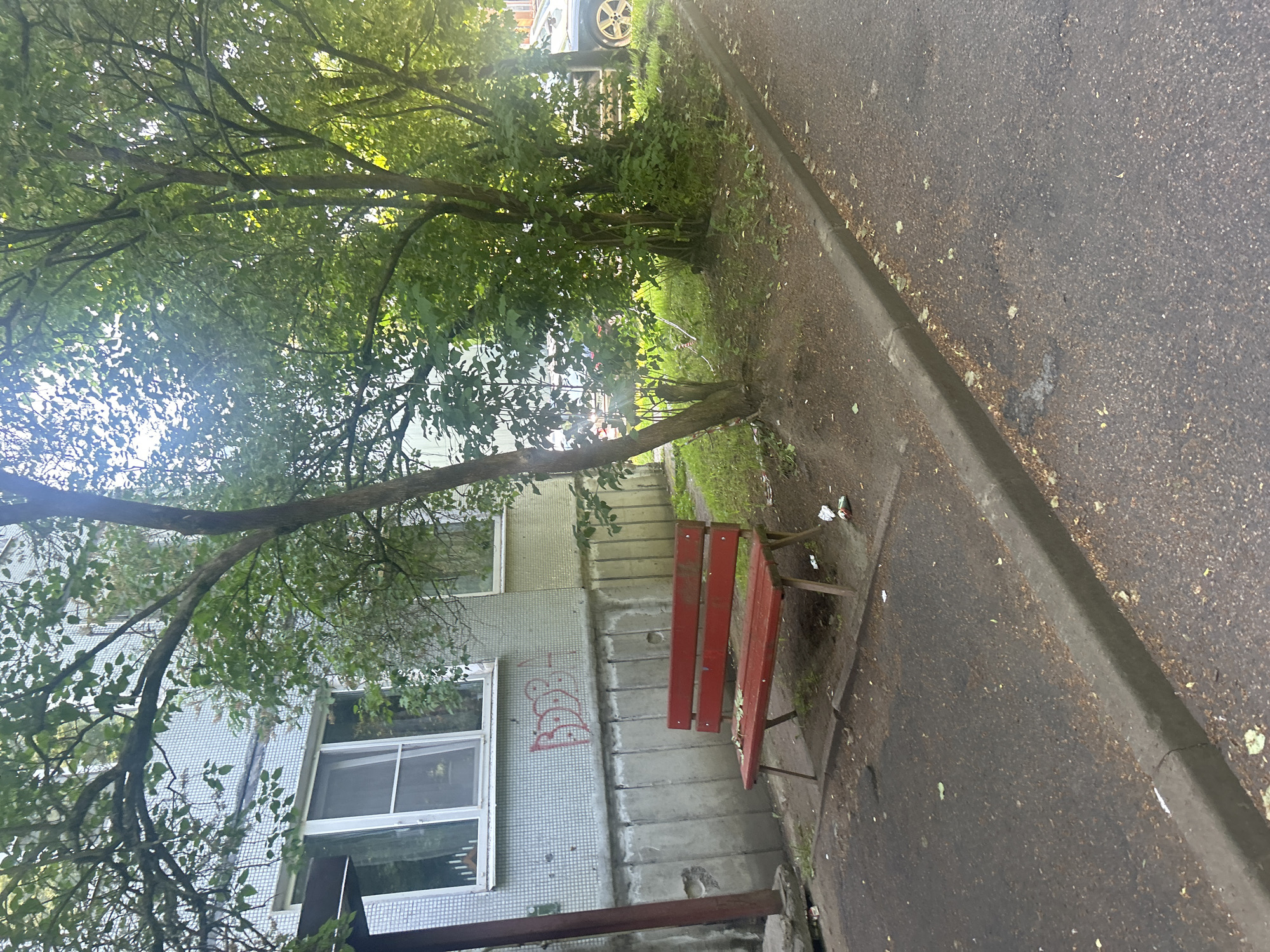 A red, weathered park bench is positioned on a paved area next to a grassy strip and a building in Posyolok Imeni Morozova, Russia. The building, likely an apartment block, has a light-colored tiled facade with a concrete foundation. Red graffiti is visible on the building's facade above the bench. Mature trees with green foliage provide shade, casting shadows on the scene. The paved area appears to be a walkway or parking area and shows signs of wear and scattered debris. In the background, a dark-colored car is partially visible. The lighting suggests it is daytime.