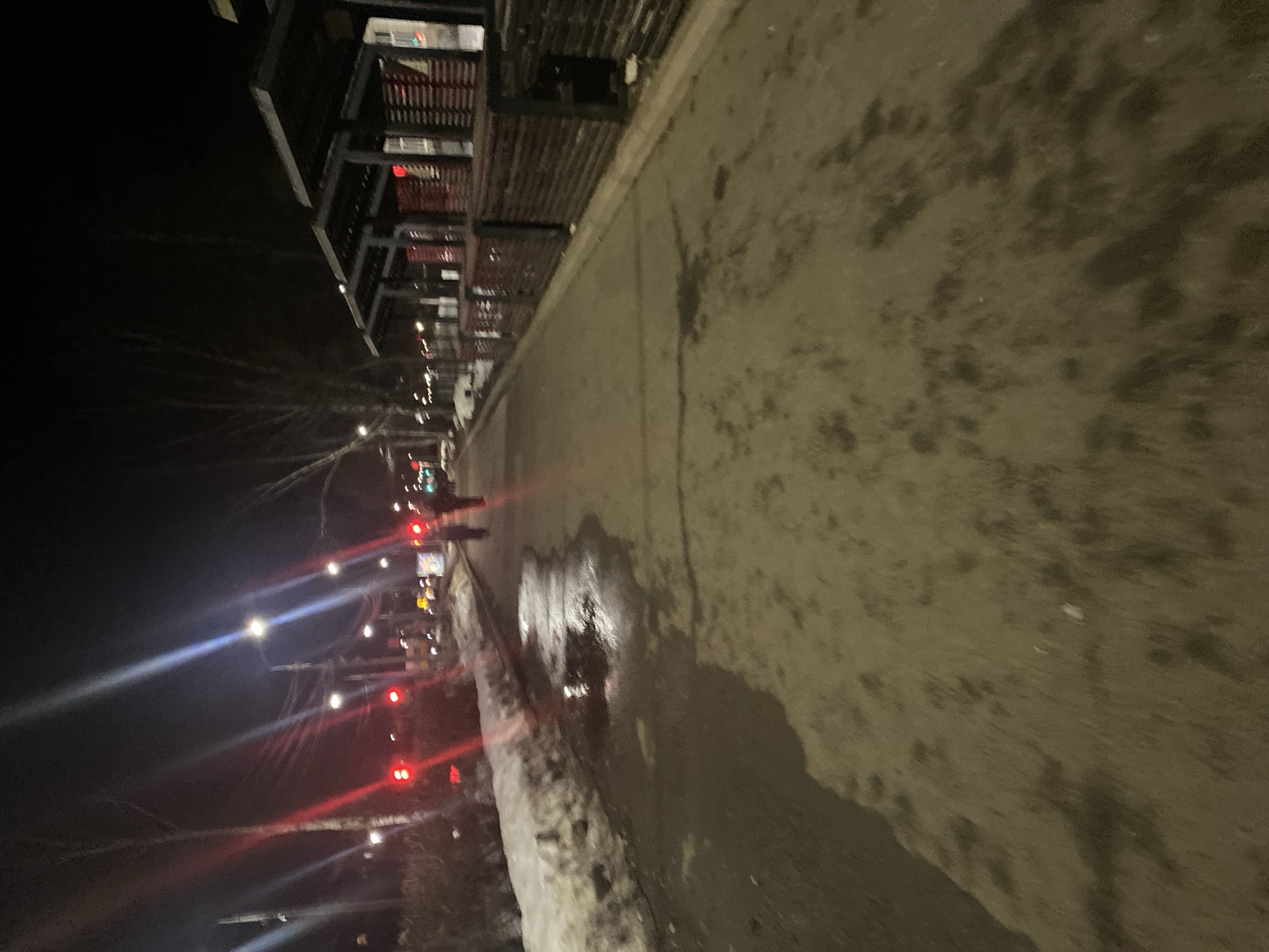 The image depicts a street scene at night in Chaikovsky, Russia. The focus is on the sidewalk, which is wet and partially covered with snow along the curb. A multi-story building with dark balconies and some illuminated windows is situated on the left side of the frame. In the background, streetlights and traffic signals are visible, suggesting a busy street with passing vehicles. A person is seen walking on the sidewalk, their silhouette illuminated by streetlights. The overall atmosphere is dark and somewhat indistinct due to the nighttime conditions.