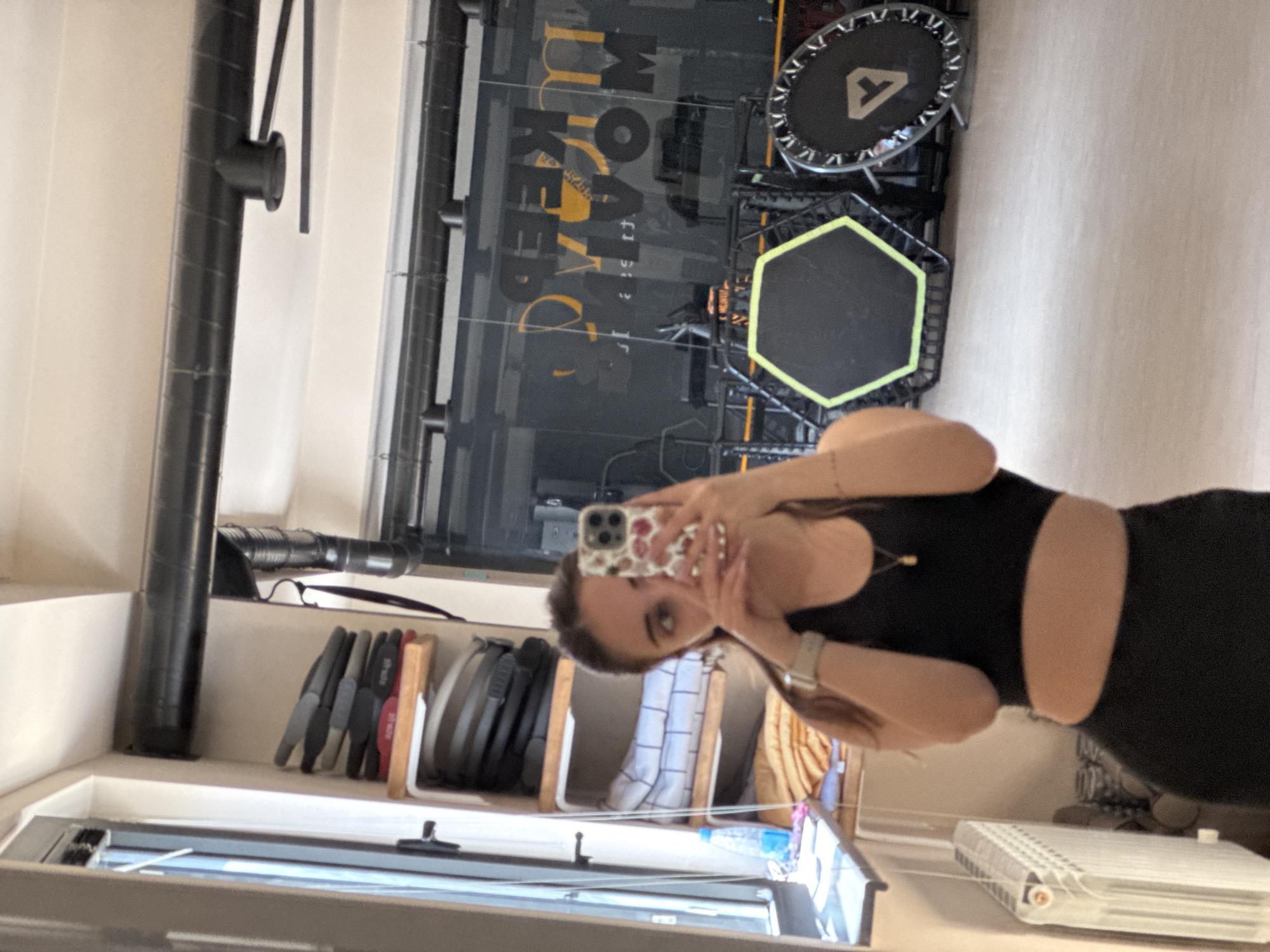 The image is a selfie taken in a mirrored space, likely a fitness studio in Yerevan, Armenia. The reflection reveals a woman holding a smartphone, possibly taking the picture. She is wearing a black workout outfit, revealing her abdomen. In the background, fitness equipment such as trampolines and weights are visible, along with a glass wall featuring what appears to be a fitness-related logo or text. The surrounding environment seems well-lit and modern.