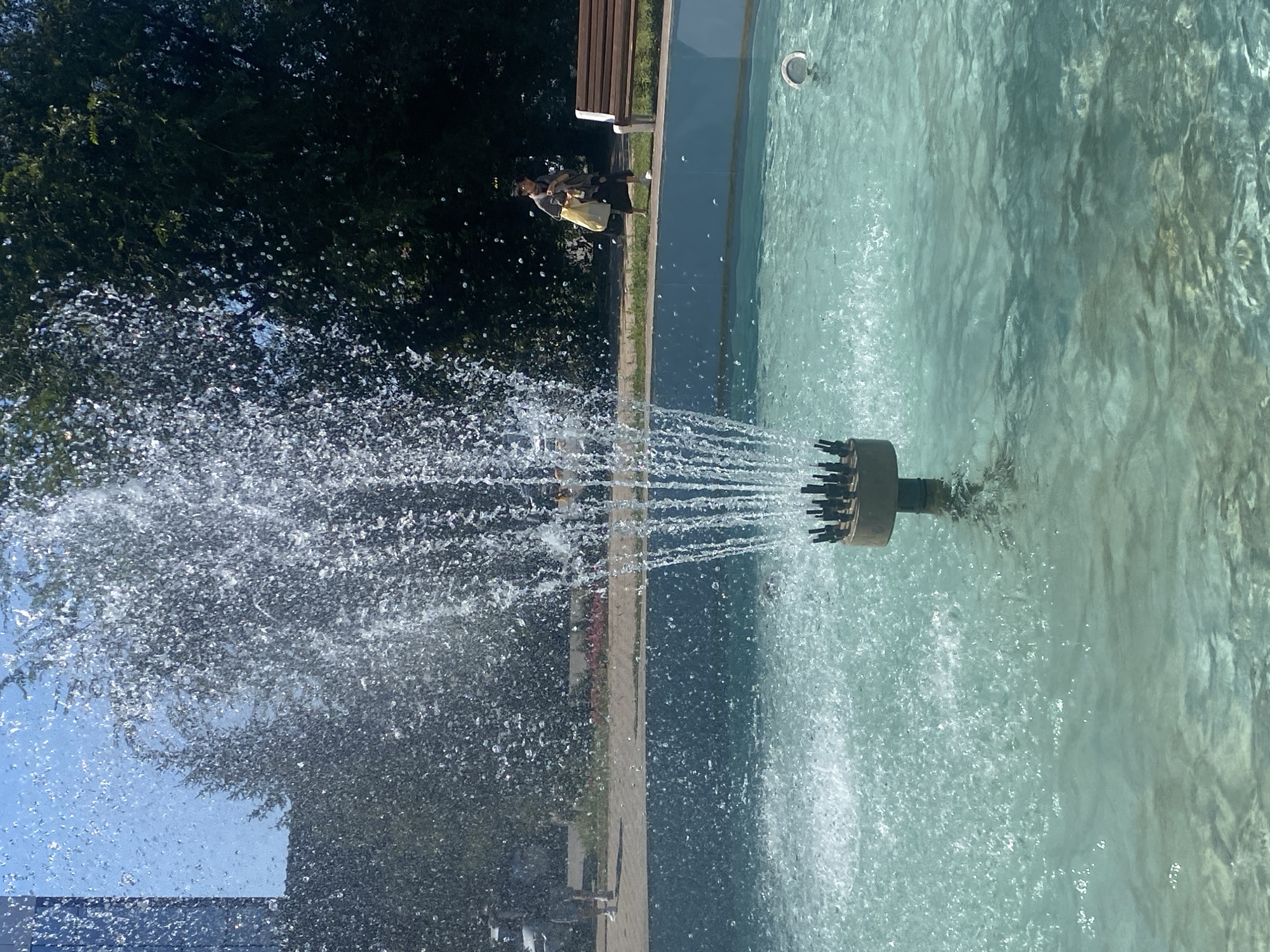 A fountain in a city park in Oral, Kazakhstan, features a central nozzle spraying water upwards and outwards. The water in the fountain's basin is a clear turquoise color. In the background, trees line the perimeter of the park, and a paved walkway is visible. Two individuals are present: one person, partially obscured by the fountain spray, stands on the walkway in the distance. Further to the right, another person walks along the walkway carrying a yellow bag, with a park bench visible nearby. The sky above is clear and blue.