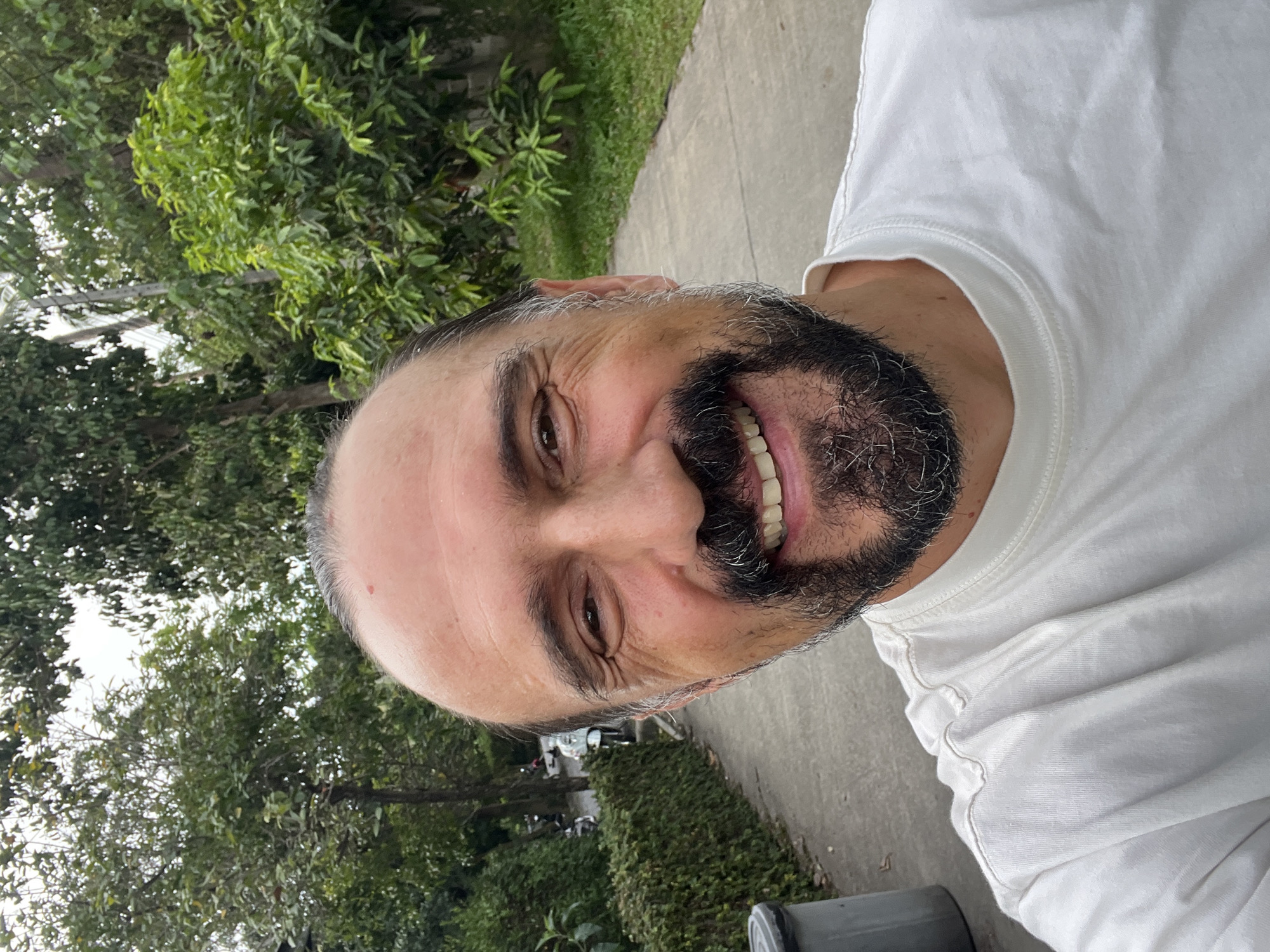 A man with a dark beard, greying hair, and fair skin is smiling at the camera while standing outside in what appears to be บ้านบางรักษ์, Thailand. He is wearing a white t-shirt. Behind him is a sidewalk, green bushes, and trees. Cars are visible parked in the background.