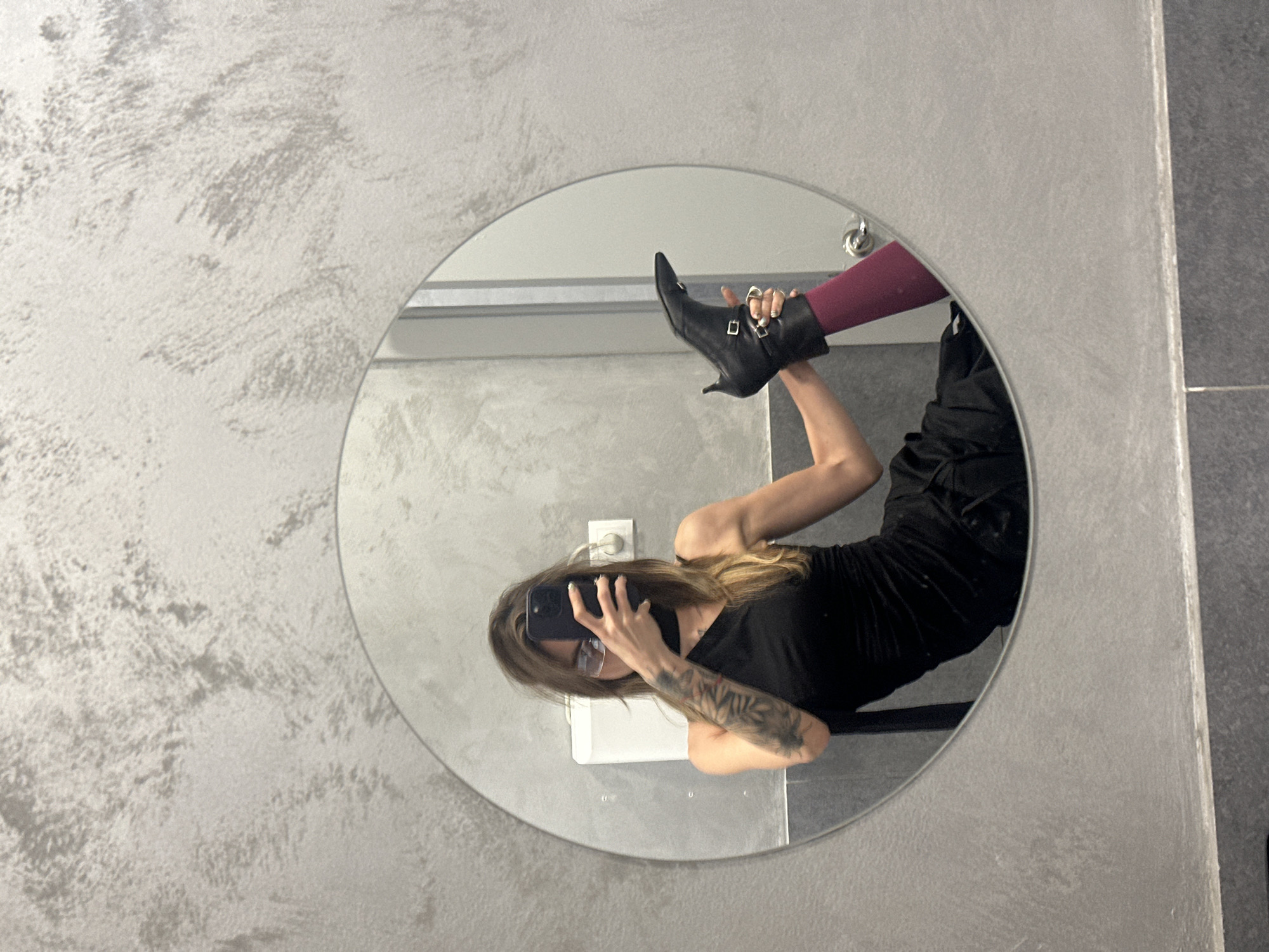 A person is reflected in a round mirror in what appears to be a restroom in Moscow, Russia. The person is wearing a black one-shoulder dress, dark tights, and black heeled boots with buckle details. They are holding up one leg, showcasing the boot and tights. The wall behind the mirror has a textured, grey finish. A white dispenser is visible on the wall to the left of the mirror, and what appears to be an electrical outlet is to the right of the dispenser.