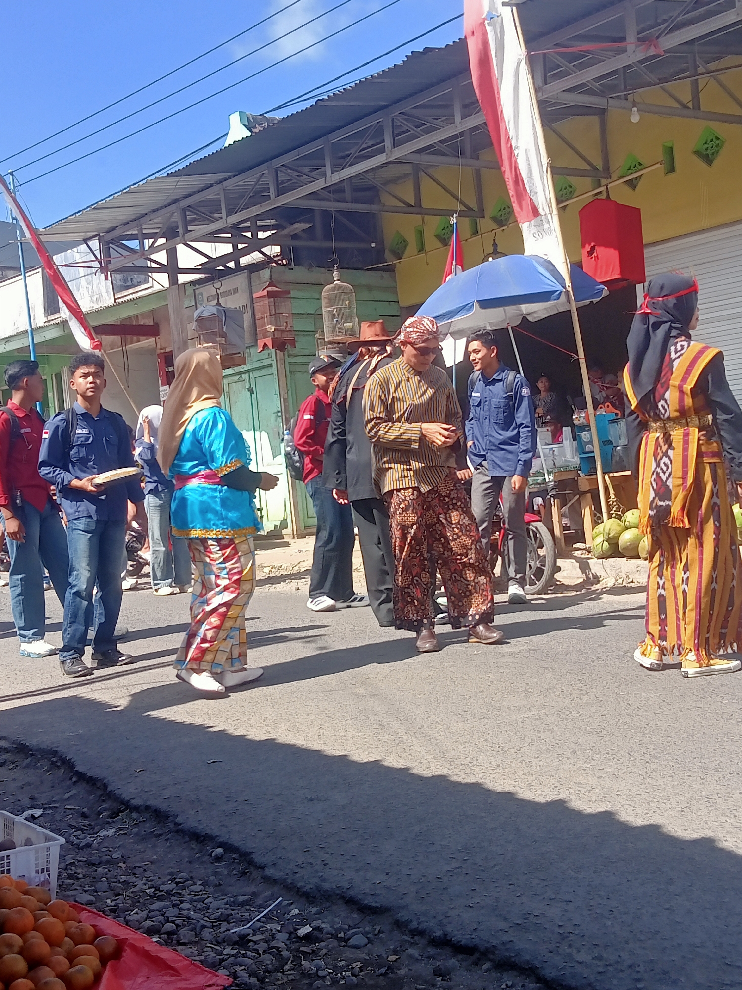 The image depicts an outdoor scene in Pule, Indonesia, likely during a cultural event or parade. Several individuals are present on a paved street.
In the foreground, on the right side, a woman wearing a traditional yellow and orange patterned dress and a black headscarf with a red band is standing. She is facing away from the camera. To her left, a man in a patterned brown and yellow striped long-sleeved shirt and brown patterned pants is walking and looking towards the left. He is wearing a headscarf.
Further to the left, a young man in a blue uniform shirt and blue jeans is holding a flat, light-colored object. He is looking forward. Beside him, to his left, another young man in a red shirt and blue jeans is walking.
Behind these individuals, a building with a corrugated metal roof and green siding is visible. A red structure, possibly a speaker or decoration, is mounted on the building. A blue umbrella is set up near a market stall. Birdcages are hanging from the building. To the far right, a pile of green, round objects, likely coconuts, are stacked. In the lower left corner, a red tarp is spread out with a white plastic basket and a pile of orange fruits, likely oranges, arranged on it.
The sky is clear and blue, indicating a sunny day. The overall atmosphere appears to be lively and communal.