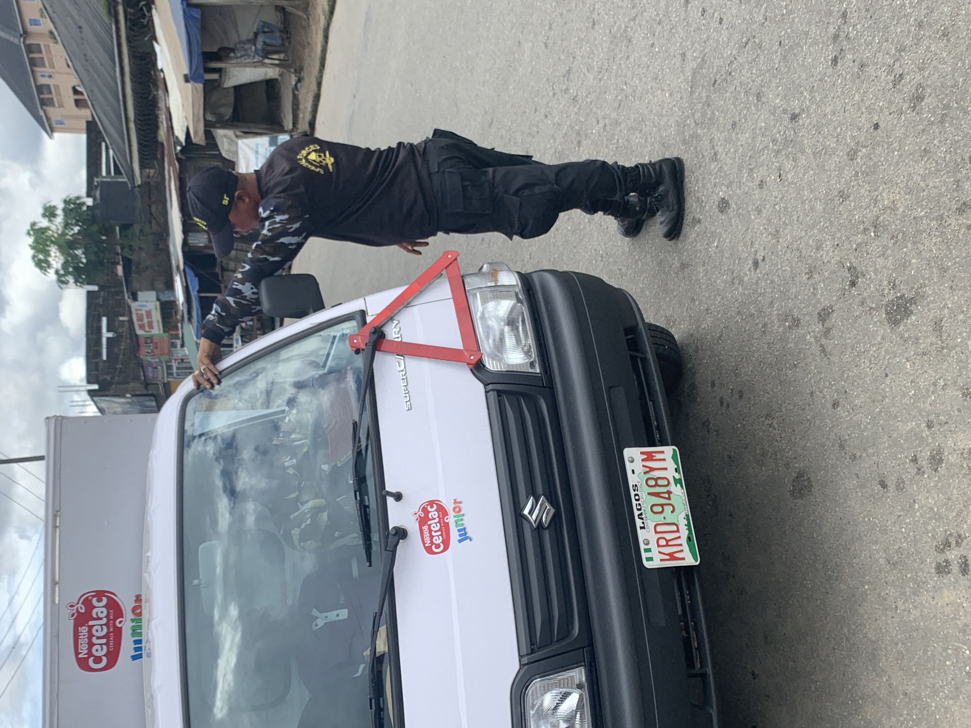A man in a black uniform with camouflage sleeves and a black cap, possibly a security guard, is standing next to a white delivery van with "Nestle Cerelac Junior" branding. He is holding a red triangular warning sign and appears to be placing it on the windshield of the van. The van has a Lagos license plate that reads "KRD-948YM". In the background, there are shops with corrugated iron roofs and a tree. The scene is on a paved street, suggesting an urban or semi-urban environment, consistent with a city like Umuode, Nigeria, though the specific location cannot be definitively confirmed from the image alone. The sky is cloudy.
