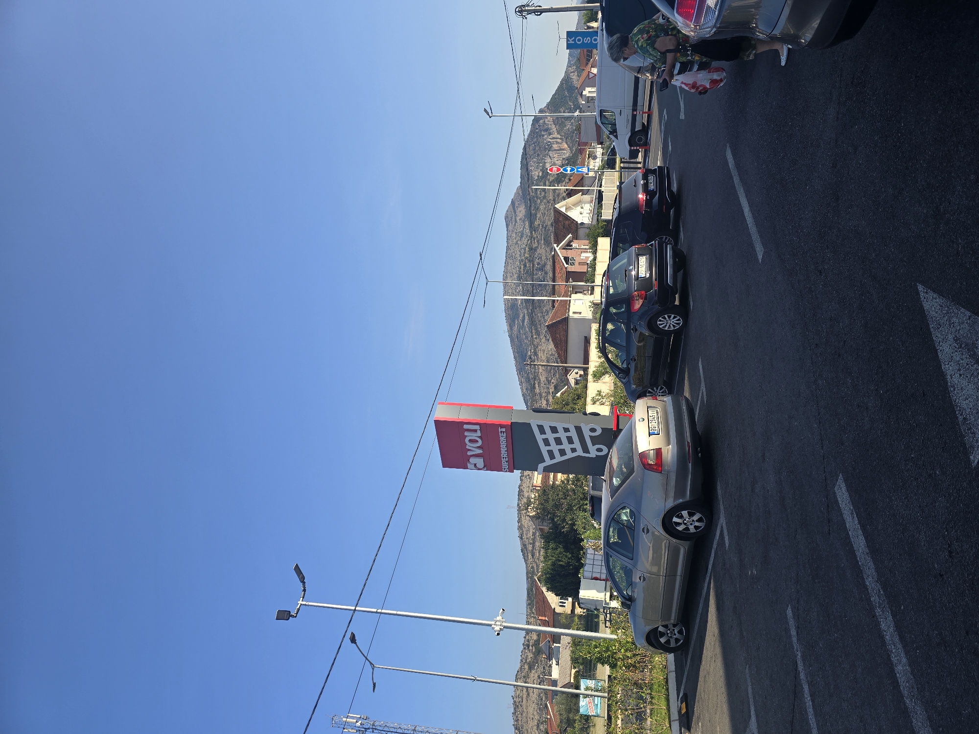 The image depicts an outdoor scene in a city, likely Podgorica, Montenegro, showing a parking lot in front of a supermarket. The sky is clear and blue, with a few power lines and streetlights visible. A large red and grey Voli supermarket sign stands prominently in the mid-ground. Several cars are parked in designated spots. In the background, there are residential buildings and a hilly landscape. A woman is seen walking in the parking lot, carrying a shopping bag. The overall impression is a typical day in a commercial area.