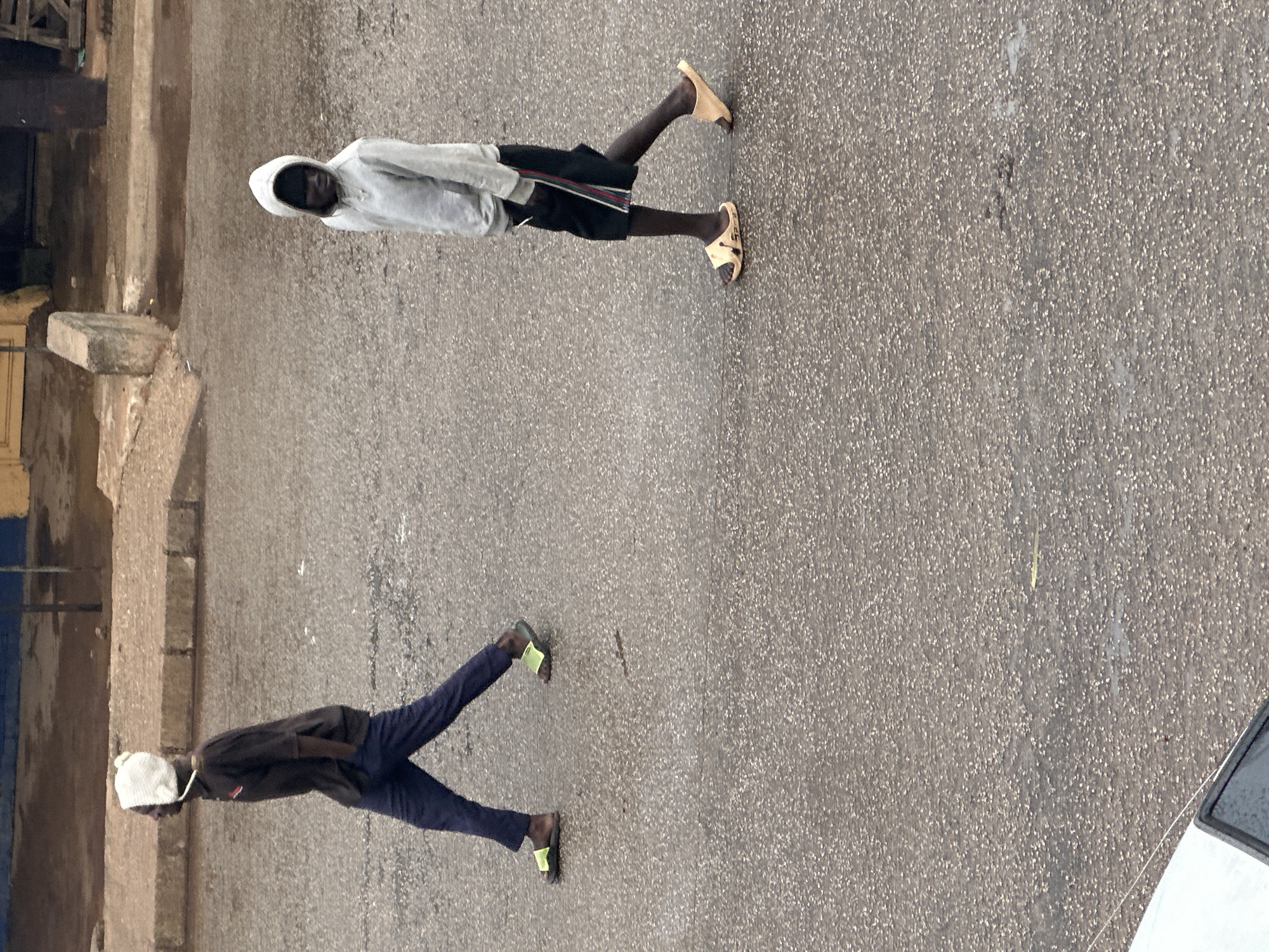 This is an aerial, medium shot of two young Black men walking on a wet, asphalt road. The man on the left, wearing a dark t-shirt and navy pants, has his hands in his pockets and is wearing a white beanie. He has yellow slides on his feet. The man on the right, wearing a grey hooded sweatshirt and black shorts with stripes on the side, is walking with his hands at his sides and is wearing tan slides. Both men are walking away from the camera towards the left of the frame. In the background, there is a curb and a building with yellow and blue paint. The road is reflective, indicating it is wet from recent rain. The scene appears to be located in Bukuru, Nigeria.