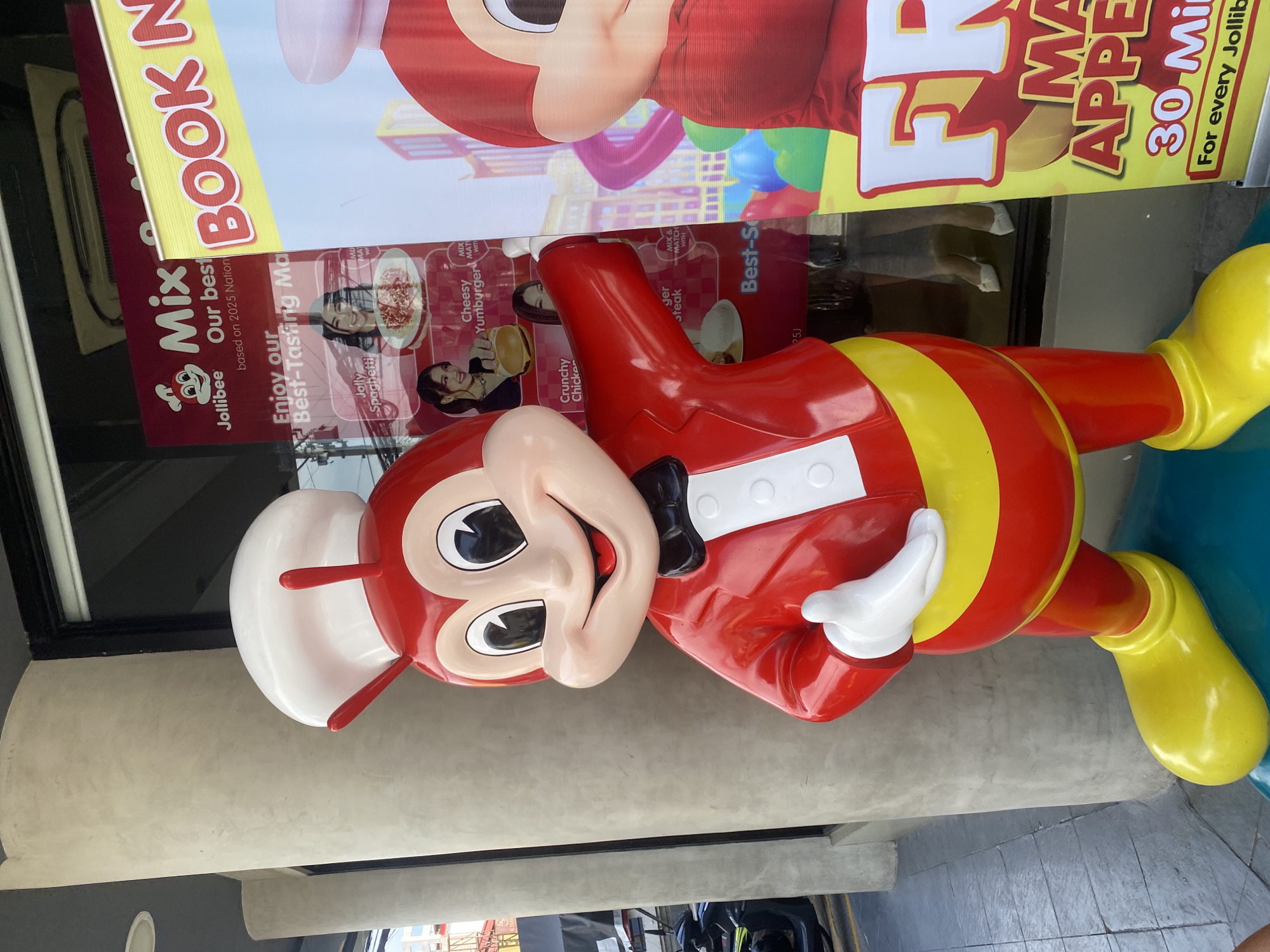 A vibrant, life-sized statue of Jollibee, the mascot for the Jollibee fast-food chain, stands prominently in an outdoor area of a Jollibee restaurant, likely in Davao City, Philippines. The cheerful mascot, dressed in a red suit with a white shirt, black bowtie, and white chef's hat, features yellow striped gloves and boots, and holds its hands out as if greeting customers. Its large, friendly smile and wide eyes are characteristic.
Behind the mascot, a large glass window or display case is covered with multiple promotional banners and posters. The lighting suggests it is daytime. Reflections of what appear to be people's legs and shoes are visible in the lower part of the glass.
Visible text on the banners includes:
* "Mix" with the Jollibee logo.
* "Our best based on 2025 Nation".
* "Enjoy our Best-Tasting Ma".
* "BOOK N".
* Images of food items like "Jolly Spaghetti", "Crunchy Chicken", "Cheesy Yumburger", and "Burger Steak" are displayed, often alongside smiling individuals.
* Phrases like "MIX & MATCH WITH" are repeated.
* Further text on a right-side banner reads "FR" (likely part of "FREE"), "MA APPE" (possibly "MADE HAPPY" or "MAGIC APPEAL"), "30 Min", and "For every Jollib" (likely "For every Jollibee").
* A small code "25J" and "MD MA" are also visible.
The scene captures a typical entrance to a Jollibee branch, designed to attract customers with its iconic mascot and appetizing food advertisements.