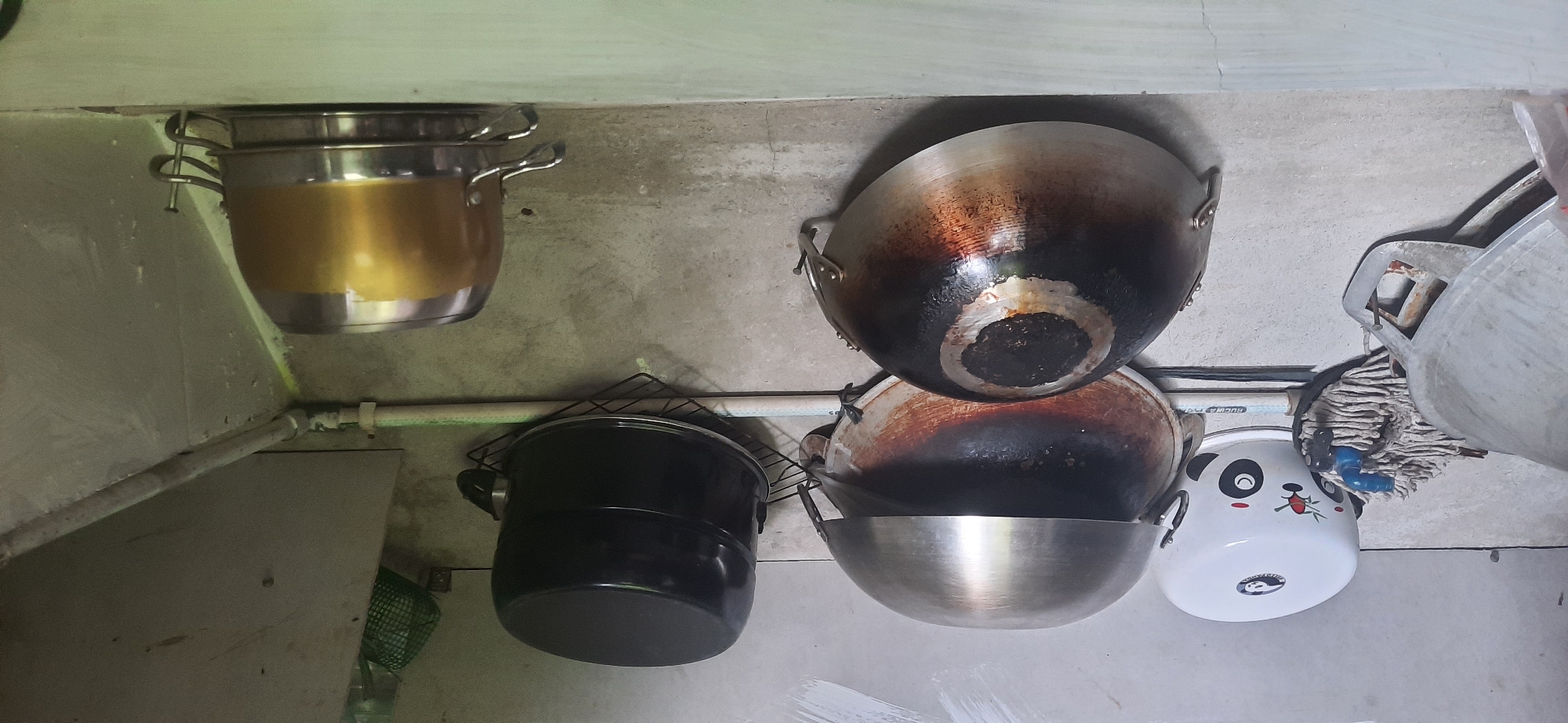 The image depicts a collection of cooking pots hanging on a wall, likely in a kitchen area. In the upper right, a large golden pot is suspended. Below it and to the left, a black pot is mounted on a wire rack. Further down and to the left, two large, well-used woks are hanging, showing signs of significant heat and use. To the left of the woks, a small white pot with a panda design is visible. Beneath these pots, a mop head with a blue faucet is resting against the wall. In the lower right, a portion of what appears to be a gas tank or similar container is visible. The wall itself is concrete and shows signs of wear and tear, with some discoloration and cracking. A white pipe runs vertically along the wall, suggesting it might be a water pipe or part of a plumbing system. The overall impression is of a functional, perhaps somewhat cluttered, kitchen space. The location is identified as Cisauk, Indonesia.