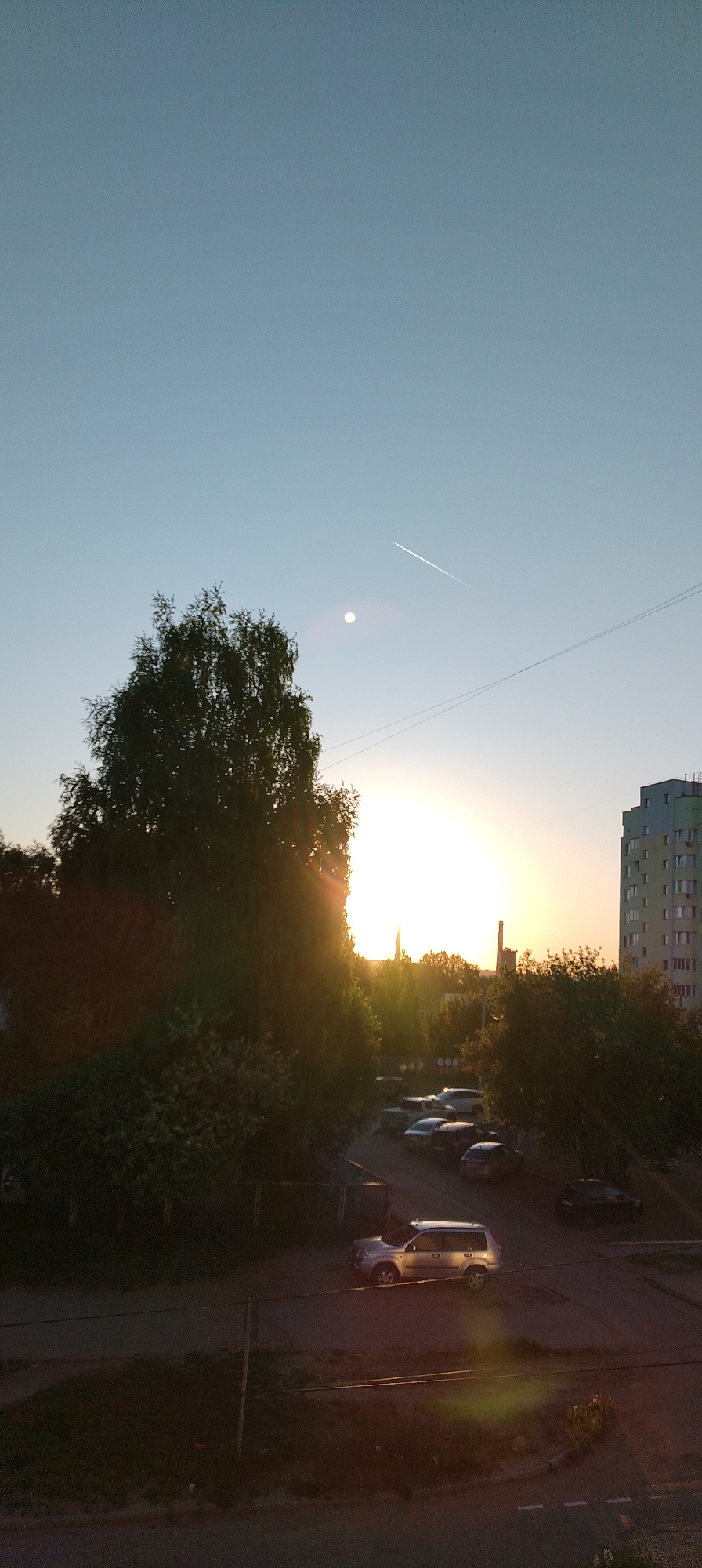 The image shows an urban scene at either sunrise or sunset. The sky is light blue, with a bright orb that is likely the sun. There is a contrail from an aircraft. A tall tree obscures a portion of the sun. Below the tree is a parking area with several cars. A tall residential building is on the right side of the image. There is no indication of people. The photograph was possibly taken in or near the city of Chaikovsky, Russia.