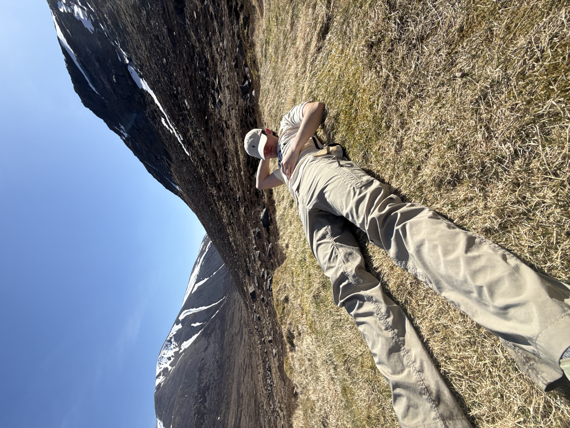 A person is lying on a grassy slope with mountains in the background, under a clear blue sky. The person is wearing a light-colored t-shirt, khaki pants, and a baseball cap, with their right hand behind their head. The grass is mostly brown with patches of green. The mountains are rocky with patches of snow. The photograph is taken from a low angle. The location appears to be a natural environment, possibly a mountainous region. Unknown, Unknown.