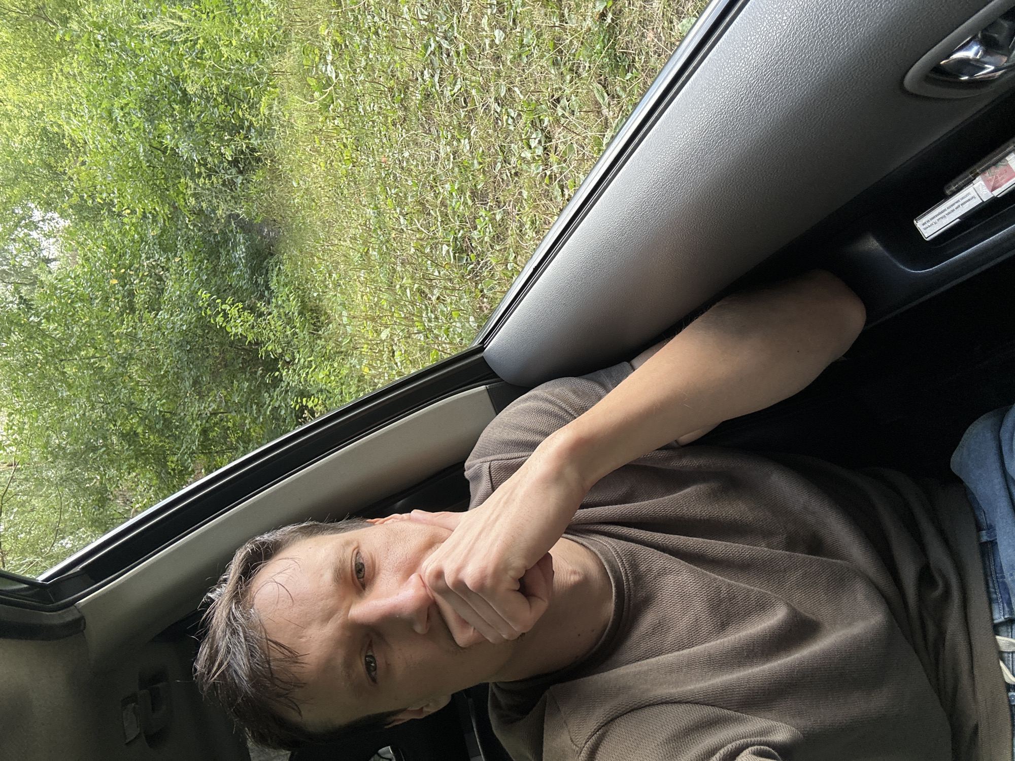 A man with short brown hair is sitting in the driver's seat of a car, looking at the camera. He is wearing a brown t-shirt and blue jeans. His right hand is resting on his chin, and his eyes are open. The car is parked on a dirt road next to a dense forest with green foliage. The sun is shining, casting a slight shadow on the man's face. In the car's door pocket, a pack of cigarettes is visible. The location is Shatava, Ukraine.