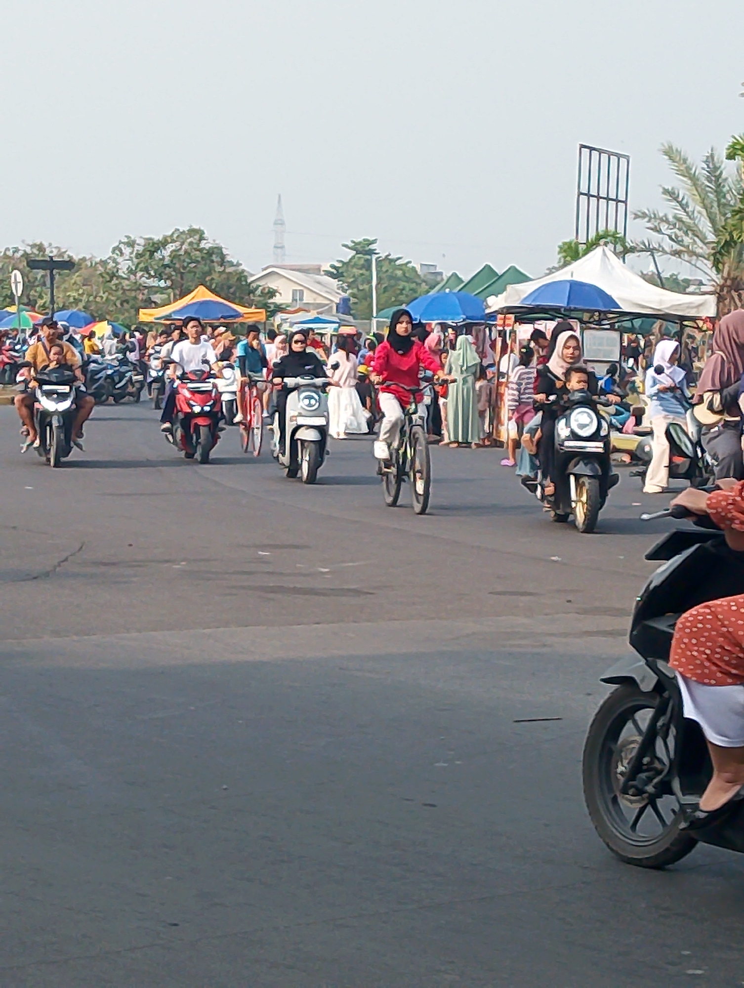 The image depicts a street scene in Karawang, Indonesia, likely a marketplace or event area, with a paved road in the foreground and a bustling crowd in the background. Several people are riding motorcycles and bicycles. On the left, a person rides a motorcycle with a child as a passenger. In the middle, another person rides a red motorcycle, and further to the right, a person rides a silver scooter, also with a child passenger. A person on a bicycle is also visible in the middle-left of the image. The background features numerous stalls with colorful tents and awnings, indicating a commercial or festive atmosphere. Trees and some buildings are visible beyond the market area. The sky is overcast and bright. The overall scene conveys a sense of lively outdoor activity.