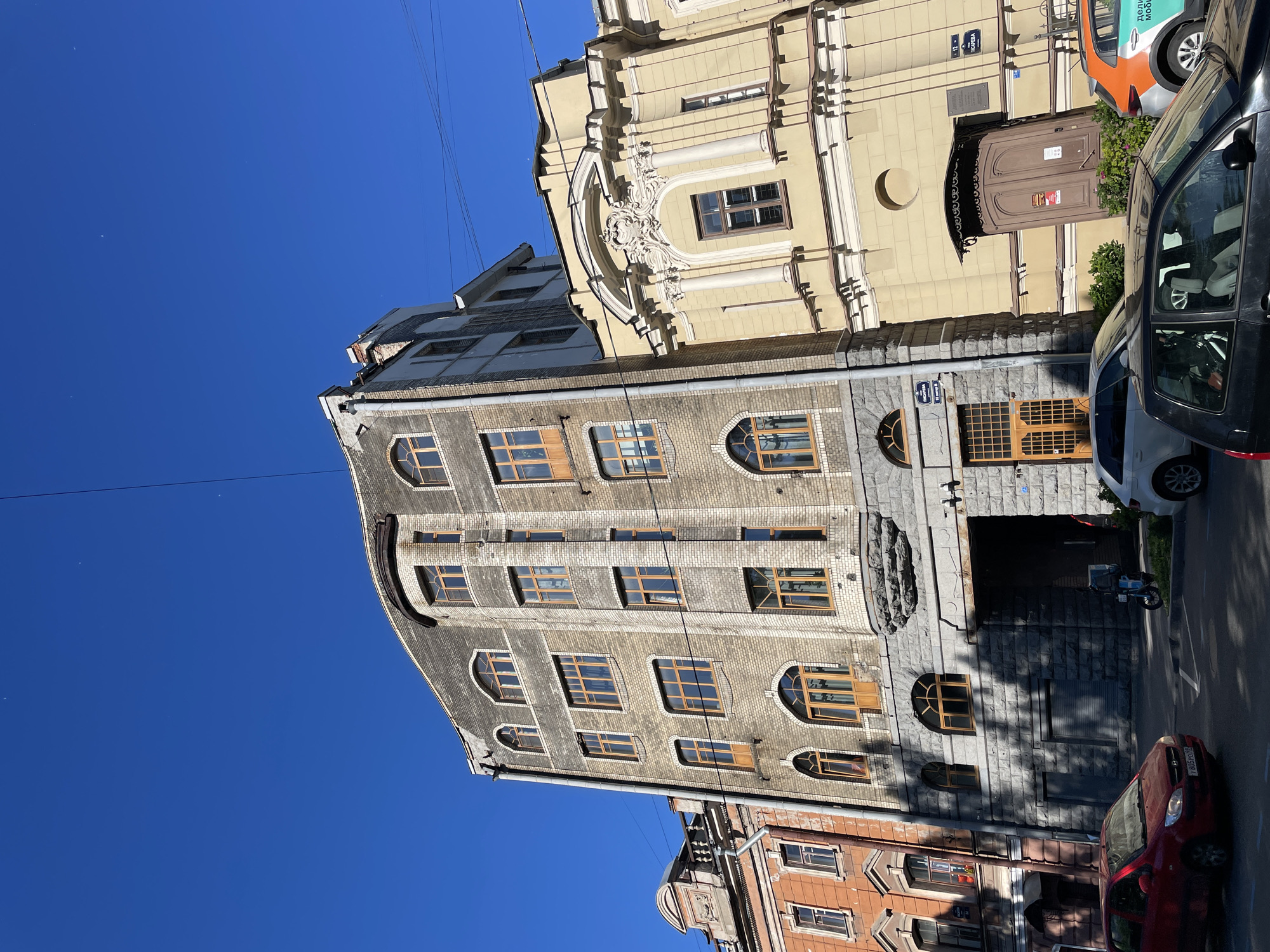 The image depicts a street scene in Saint Petersburg, Russia, featuring a mix of architectural styles under a clear blue sky. Dominating the center is a multi-story building with a curved facade, constructed from light-colored bricks with darker stone accents around the ground floor and archways. The building has numerous windows, many of which are arched and have wooden frames. To the left, a narrower building with an orange facade and decorative cornices is visible. To the right, a lighter yellow building with ornate classical detailing, including columns and a decorative pediment, stands prominently.
The street level shows parked cars, including a red hatchback in the foreground to the left, and a black hatchback further back on the right. A blue scooter with a delivery box is parked near an arched entryway of the central building. The pavement and street are visible, as are shadows cast by the buildings and trees. Power lines are strung across the sky. Street signs are visible on the buildings, with one sign on the yellow building showing "12" and Cyrillic text, and another on the central building displaying "18 Литер Б" with Cyrillic text. The overall impression is an urban environment with historic architecture bathed in bright sunlight.