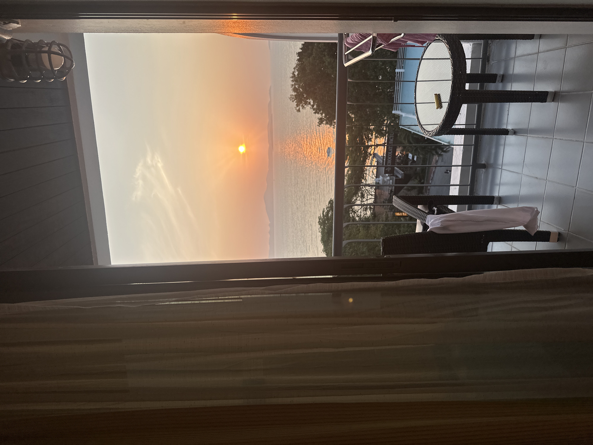 The image shows a sunset view from a balcony in Pattaya City, Thailand. The sun is setting over the ocean, casting a warm orange glow. The balcony has a small round table, a chair, and a railing overlooking the water. Trees and buildings are visible in the distance. The image is taken from inside, looking out through a doorway with curtains.