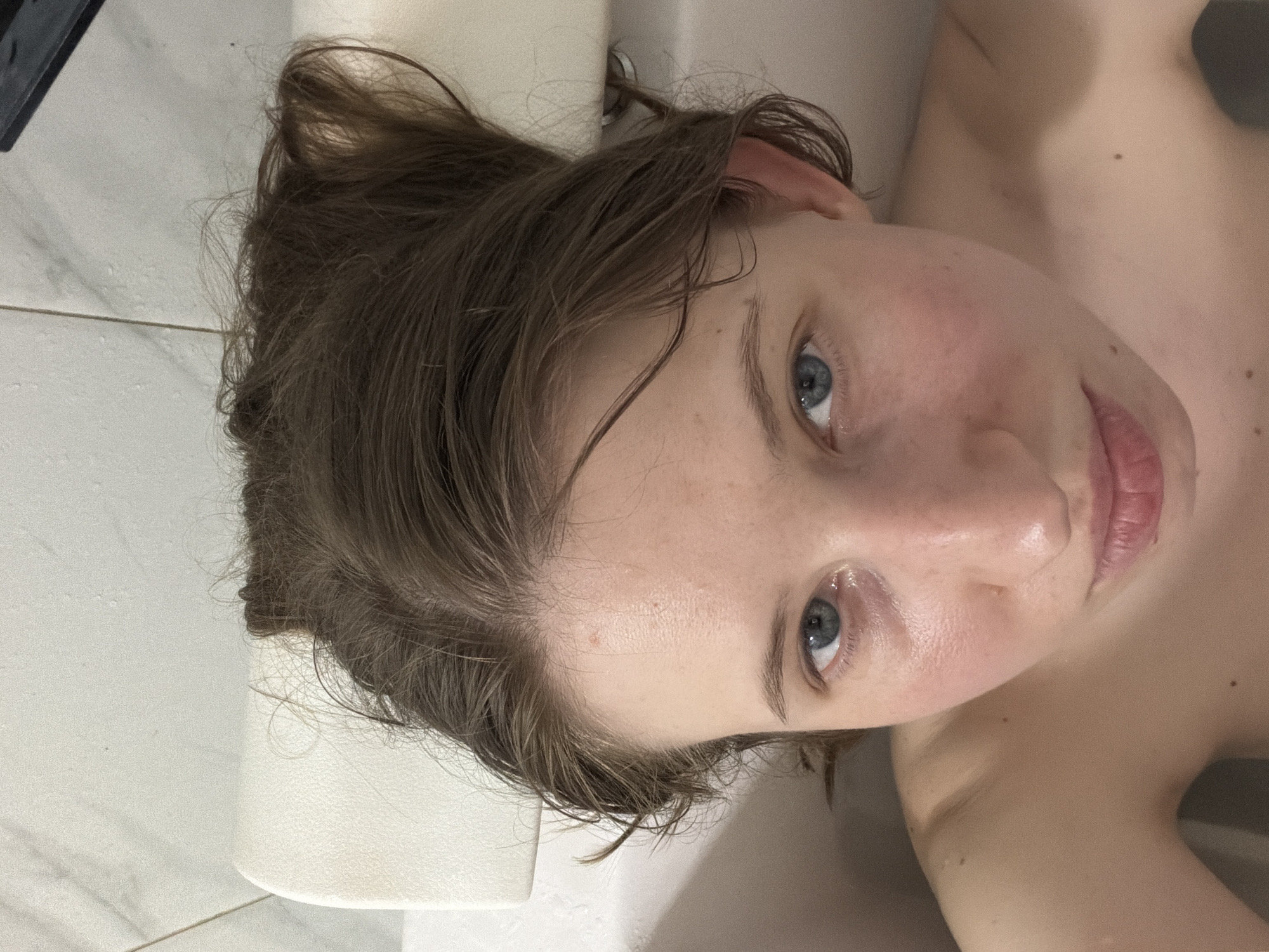 A close-up, overhead shot shows the face of a young person with wet, brown hair that is spread out on a white surface. The person has blue eyes and light skin with freckles. Their lips are slightly parted. The background is a tiled surface that appears to be in a bathroom. The image is taken from a high angle, looking down at the person. The lighting suggests it could be daytime, as there is no artificial light source visible and the natural light seems to be casting subtle shadows. The overall mood is intimate and vulnerable due to the close proximity and the person's bare skin. There is no visible text in the image.